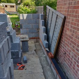 OPTIROOMS SERVICE & CONSTRUCTION SPÓŁKA Z OGRANICZONĄ ODPOWIEDZIALNOŚCIĄ - Wznoszenie muru z szarych bloczków obok ceglanej ściany. Widoczne narzędzie murarskie, ogrodzenie w tle i elementy izolacji. Prace murarskie w toku.