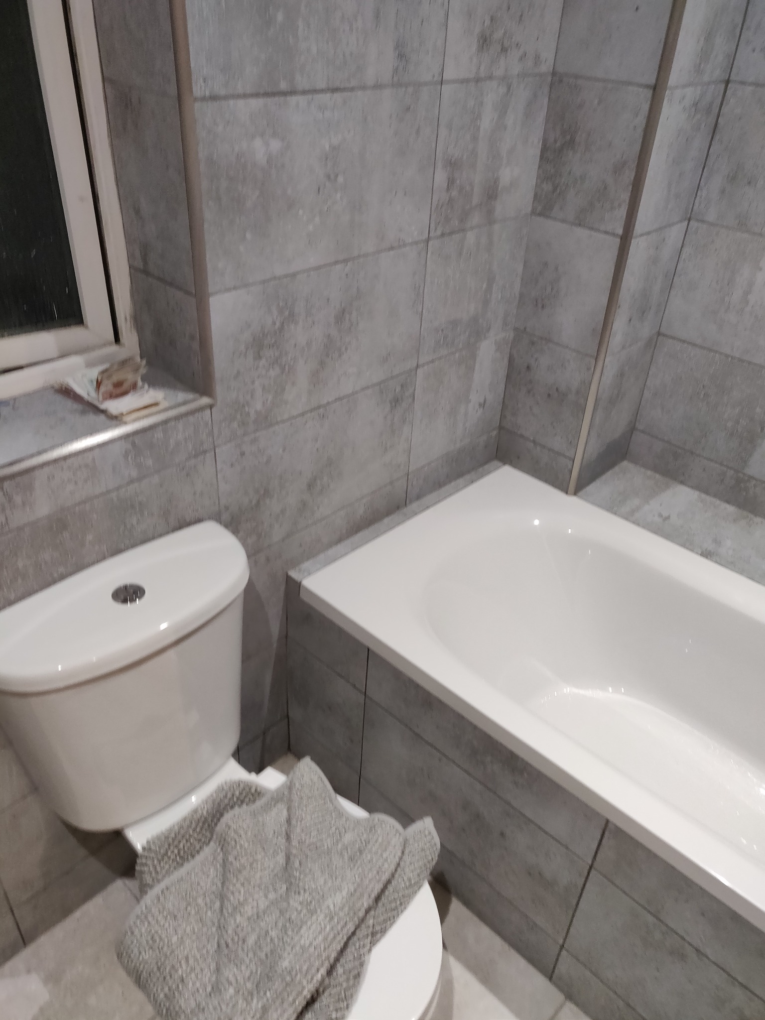 Wykończona łazienka z szarymi płytkami imitującymi drewno, białą wanną i toaletą. Na desce sedesowej leży szary ręcznik. Widok na okno z parapetem.