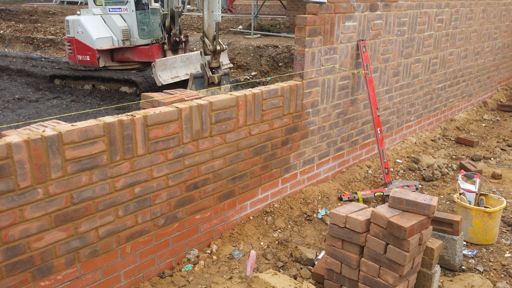 Wznoszenie muru z cegły klinkierowej z dekoracyjnym wzorem, widoczny poziomica i mini koparka w tle. Prace murarskie w toku, cegły ułożone w nietypowy wzór.