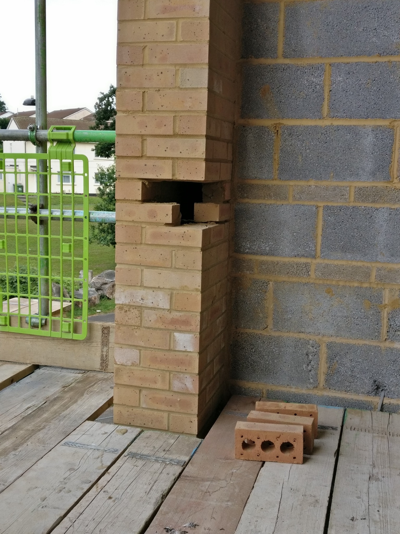 Wznoszenie ściany z cegły klinkierowej, widoczne rusztowanie z zielonymi elementami i fragment ściany z szarego betonu komórkowego. Trzy cegły na drewnianym podłożu.