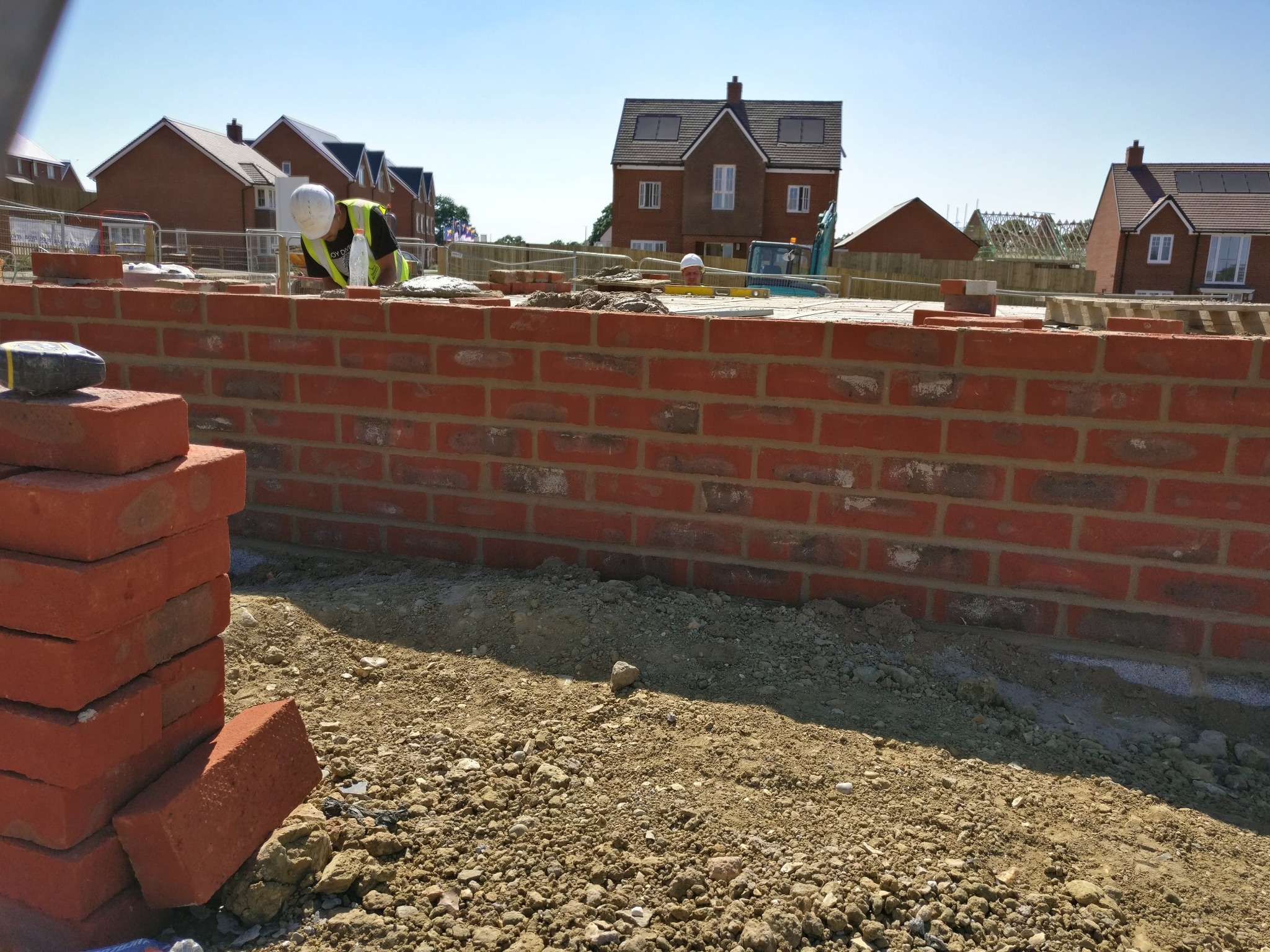 Ceglana ściana w trakcie murowania, z widocznym pracownikiem w kasku i stosem cegieł. W tle nowe domy w budowie.