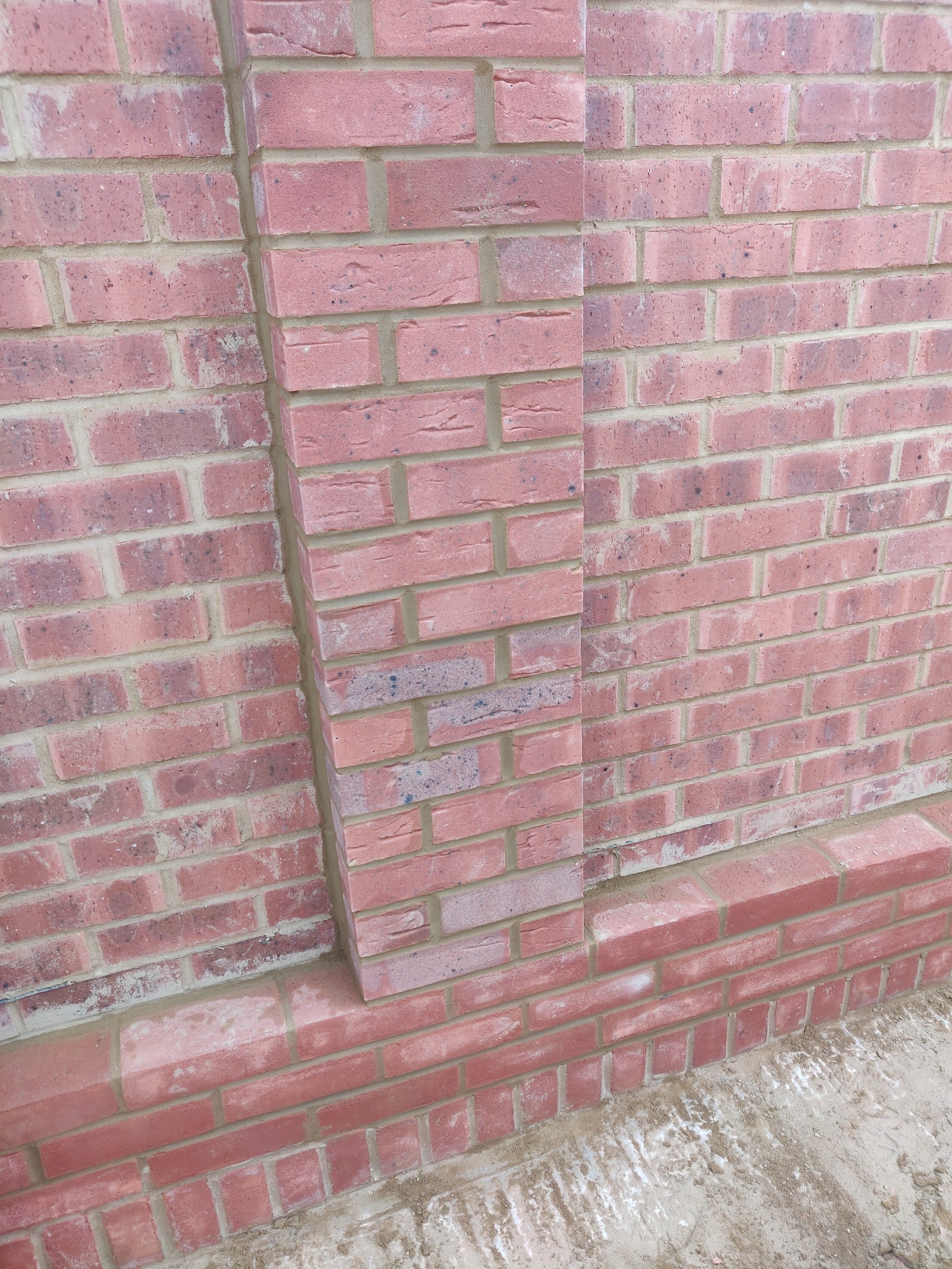 Różowa ściana z cegły z wyraźnym słupkiem wzmacniającym, widoczne spoiny, ułożona w regularny wzór, dolna część ściany z wyraźną podstawą.