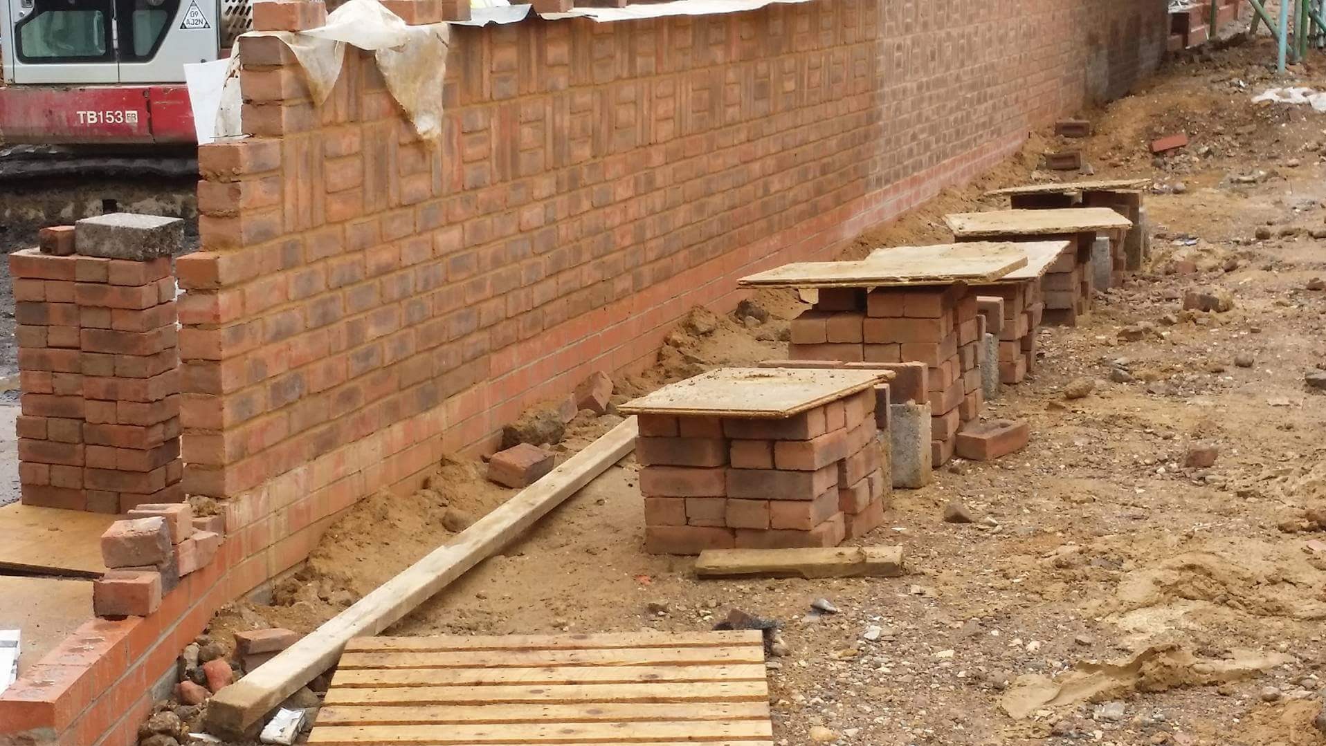 Ceglana ściana w trakcie budowy, widoczne ceglane podpory pod rusztowanie. Na drugim planie koparka.