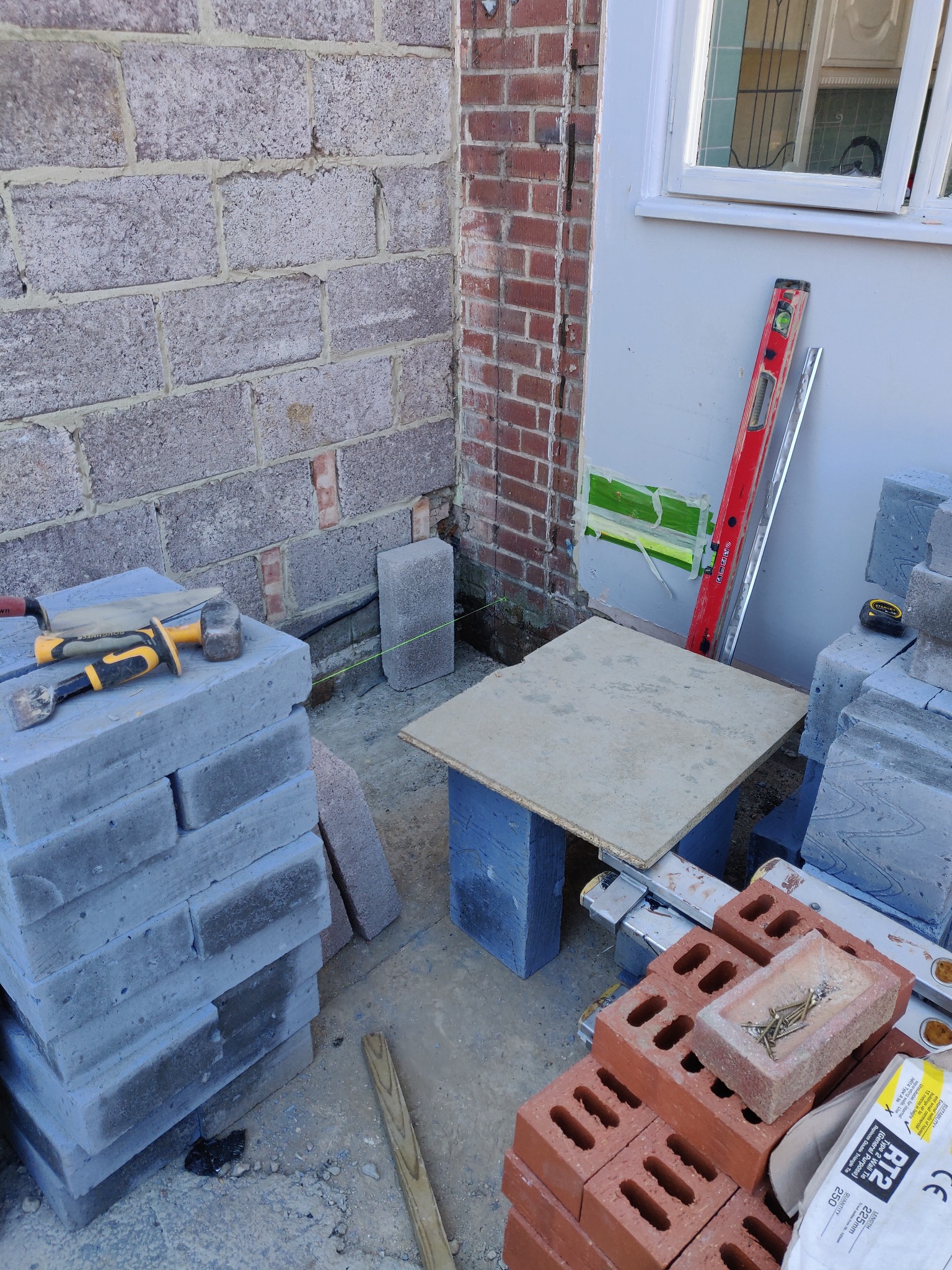 Murowanie ściany zewnętrznej z szarych bloczków, widoczne narzędzia murarskie, poziomica, cegły czerwone i biała ściana z oknem. Etap prac murarskich.