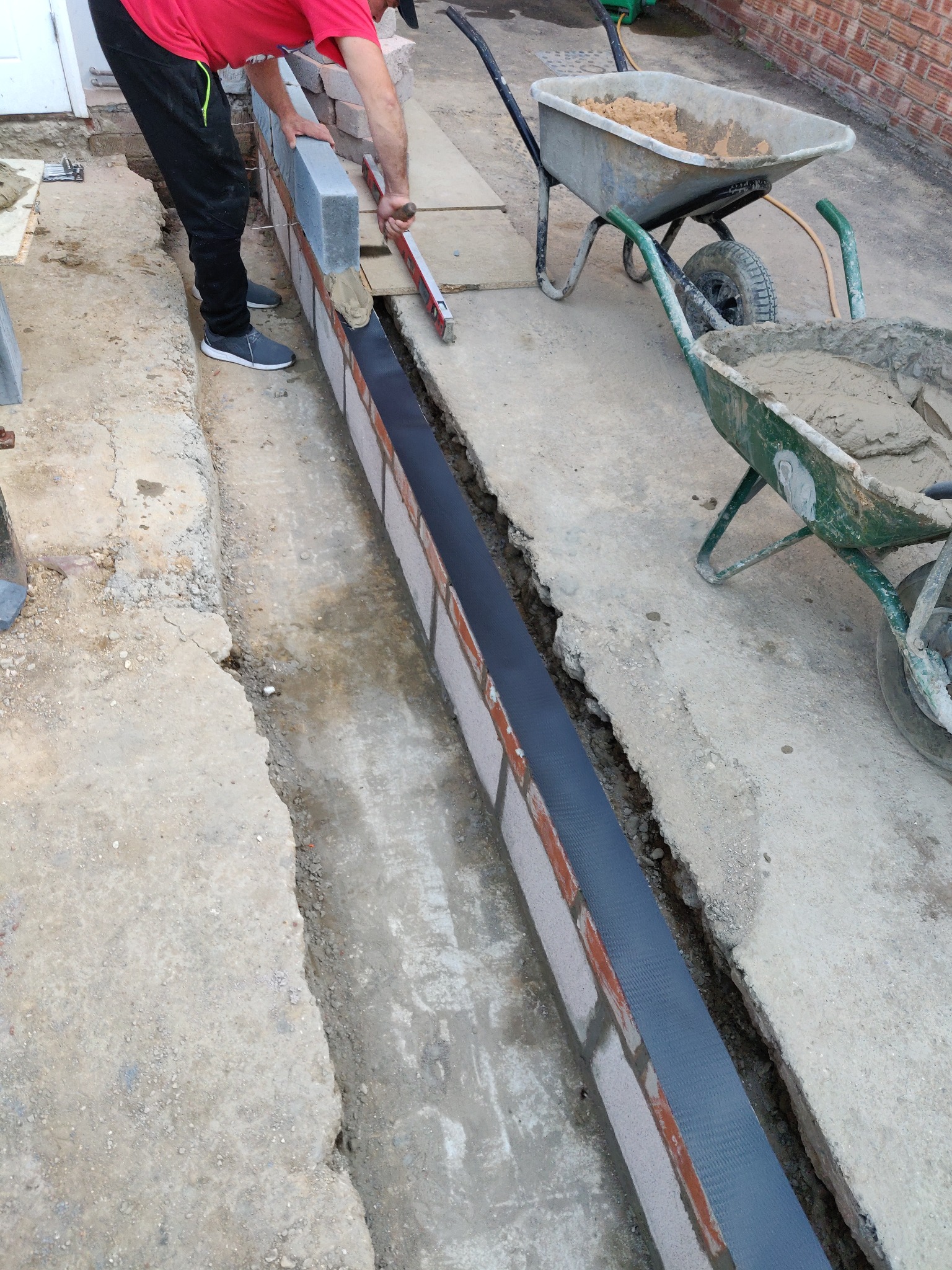 Murowanie niskiej ściany z cegieł i bloczków betonowych. Widoczny poziomica i dwie taczki z zaprawą. Czarna izolacja pionowa na fundamencie.