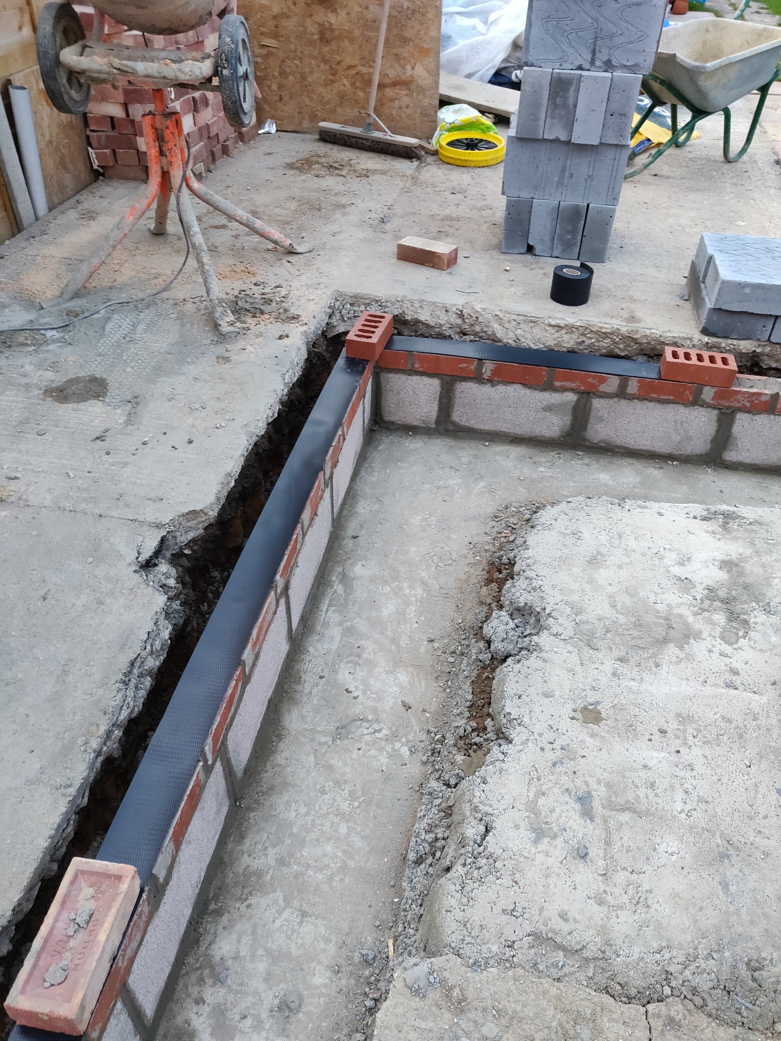 Fundament w trakcie murowania z cegły, widoczna izolacja pionowa, zaprawa i mieszalnik do betonu w tle. Fragment wykopu. Prace murarskie.