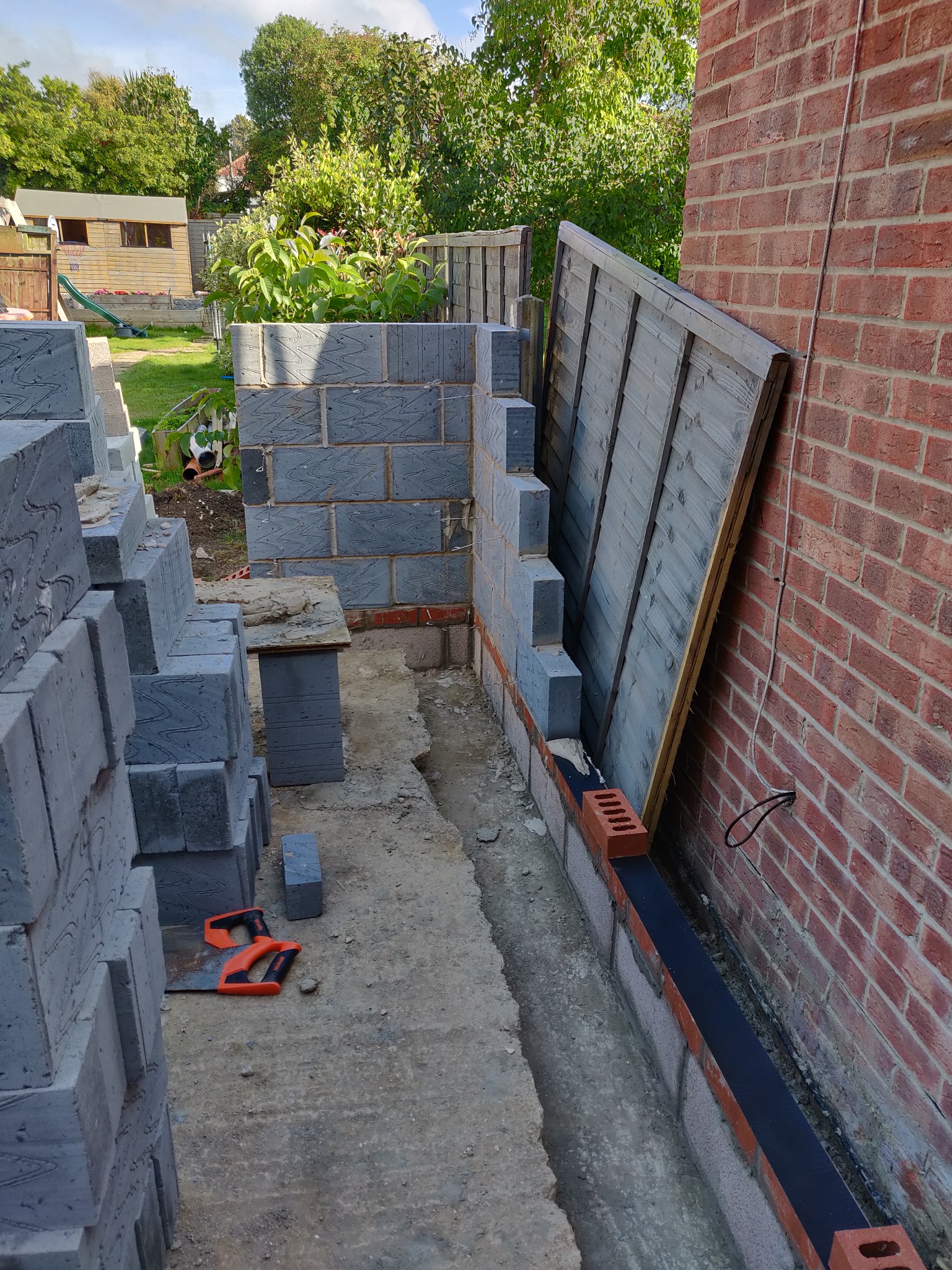 Wznoszenie muru z szarych bloczków obok ceglanej ściany. Widoczne narzędzie murarskie, ogrodzenie w tle i elementy izolacji. Prace murarskie w toku.
