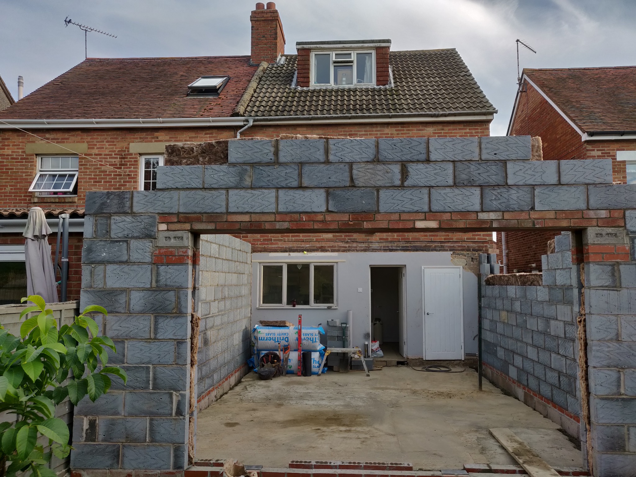 Wznoszenie ścian z szarego bloczka na fundamencie z cegły. Widoczne materiały izolacyjne i narzędzia. Budowa w toku, dom w tle.