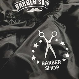 KOSZULKOTEKA - Odzież reklamowa - Nadruki - Wycena w 24h - Czarna peleryna fryzjerska z białym nadrukiem: nożyczki, gwiazdy i napis 'Barber Shop' z wąsami. Materiał lekko pognieciony, widoczne detale wykończenia.