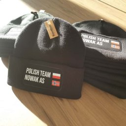 KOSZULKOTEKA - Odzież reklamowa - Nadruki - Wycena w 24h - Czarne, dziergane czapki z napisem 'POLISH TEAM NOWAK AS' i flagami Polski i Norwegii, ułożone na drewnianej powierzchni w słonecznym świetle.