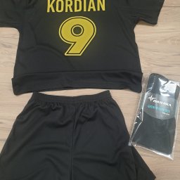 KOSZULKOTEKA - Odzież reklamowa - Nadruki - Wycena w 24h - Czarny komplet sportowy: koszulka z napisem 'POLSKA', imieniem 'KORDIAN' i numerem 9, spodenki oraz zapakowane getry piłkarskie 'ISKIERKA'.