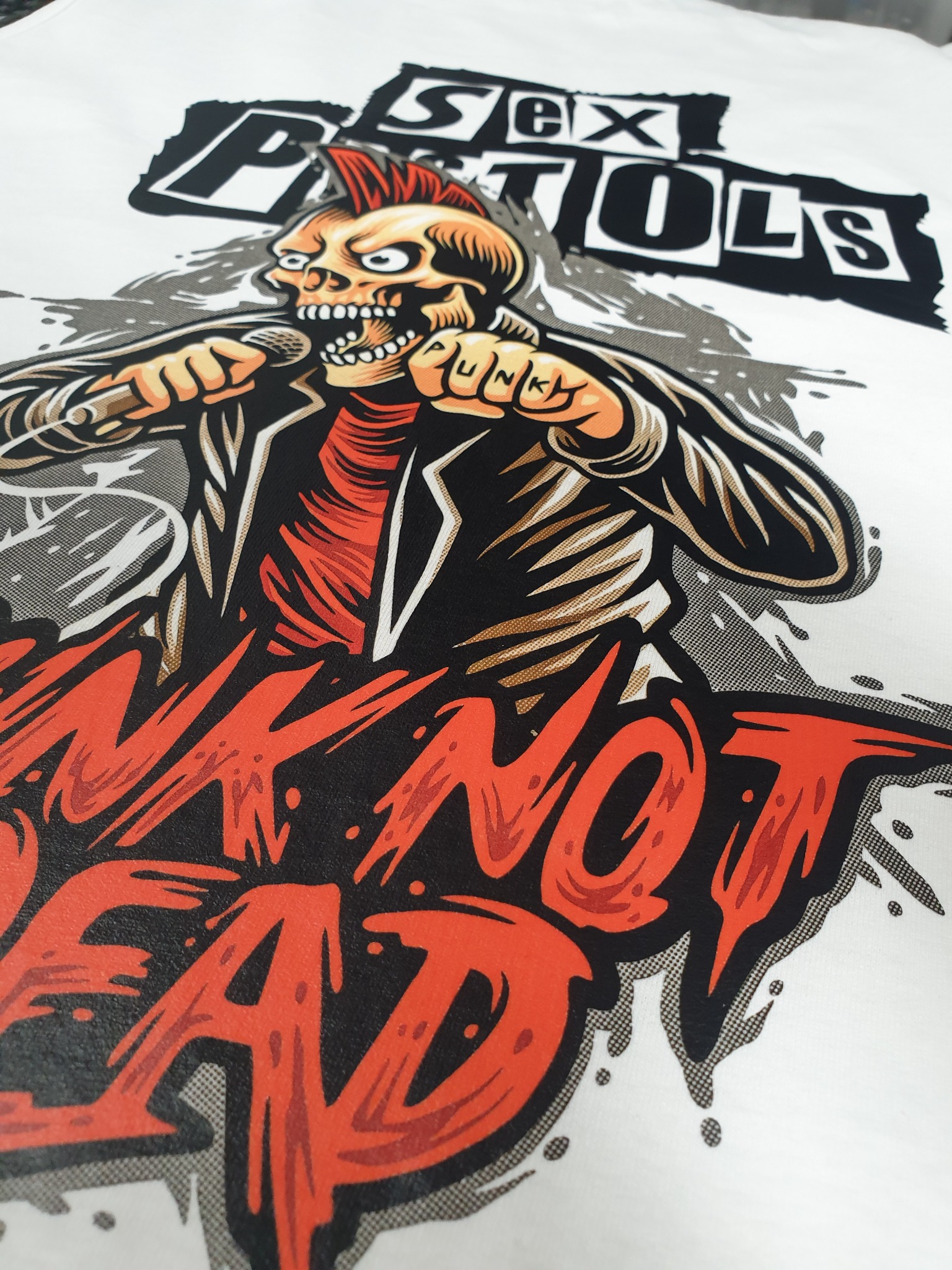 Biała koszulka z nadrukiem czaszki z irokezem, mikrofonem i napisem 'Punk Not Dead' oraz logo Sex Pistols. Wyraziste kolory, detaliczne wykonanie.