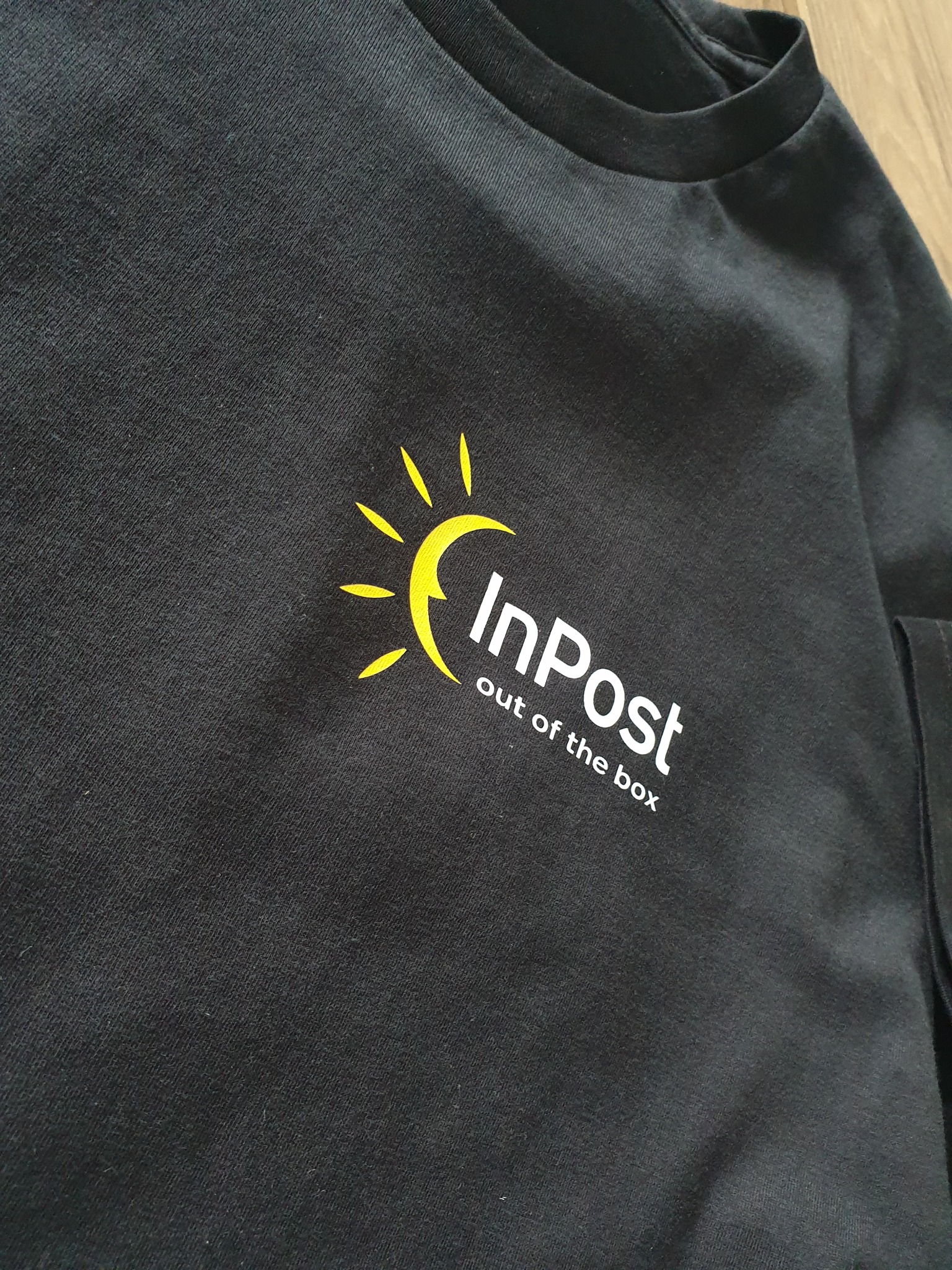 Czarna koszulka z żółto-białym nadrukiem logo firmy InPost z hasłem 'out of the box'. Widoczny fragment rękawa i kołnierzyka. Ujęcie pod kątem.