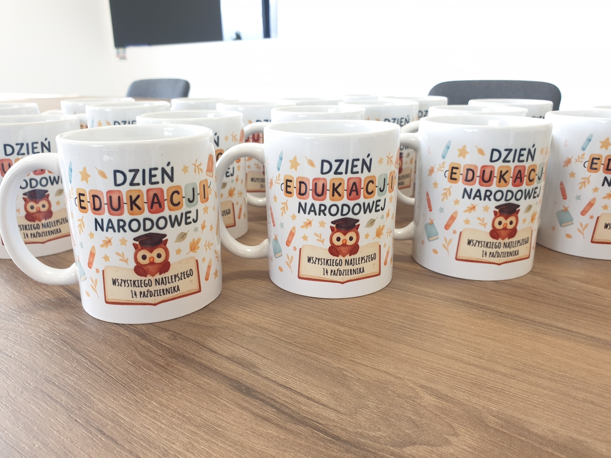 Białe kubki z nadrukiem 'Dzień Edukacji Narodowej' i motywem sowy, ustawione rzędami na stole w Świebodzicach. Idealne jako prezent dla nauczycieli.