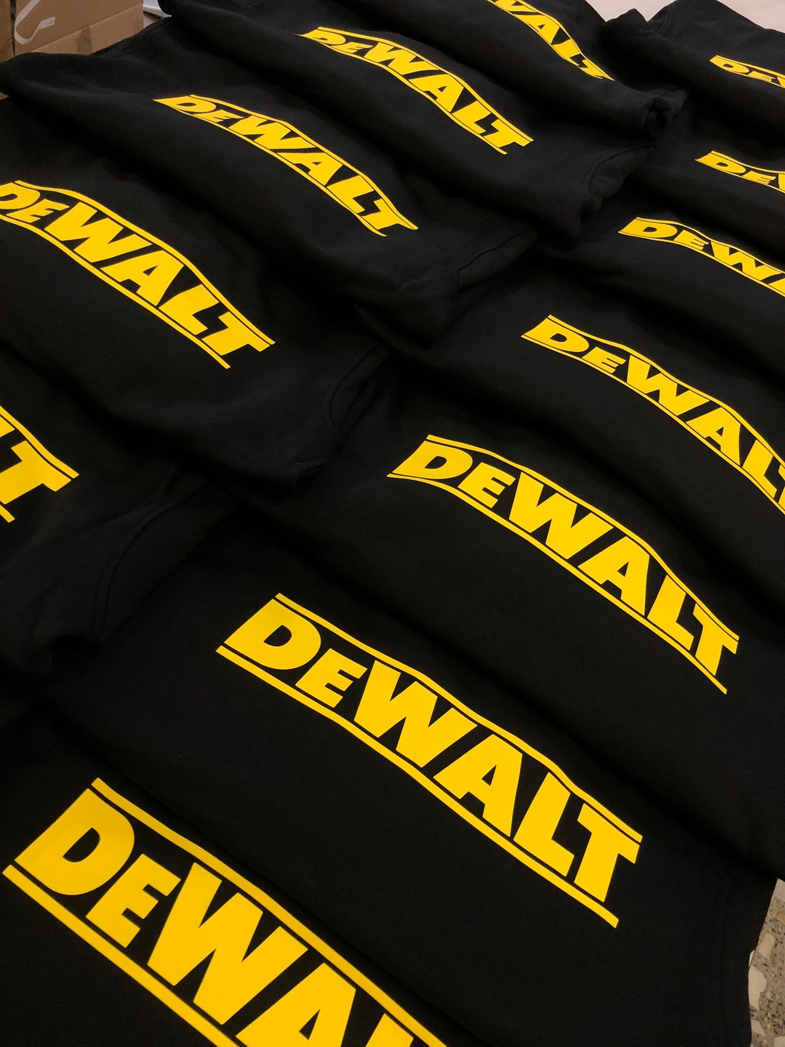 Stos czarnych bluz z żółtym, wyrazistym nadrukiem 'DEWALT'. Równomierne oświetlenie podkreśla detale logo. Ułożone w stos, gotowe do dystrybucji.