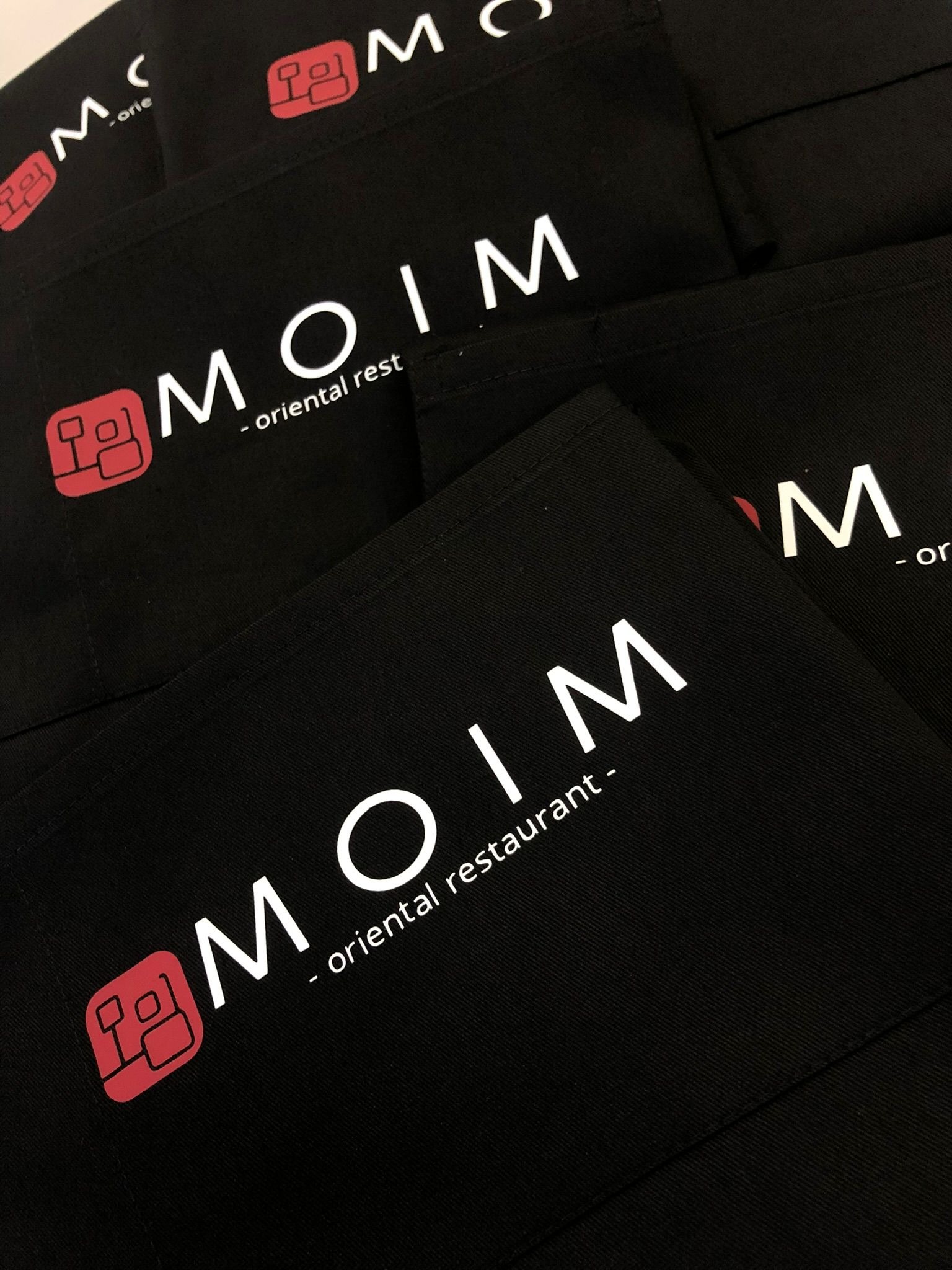 Czarne fartuchy z białym nadrukiem 'MOIM - oriental restaurant' i czerwonym logo, ułożone warstwowo, tworząc powtarzalny wzór. Detal odzieży roboczej z bliska.