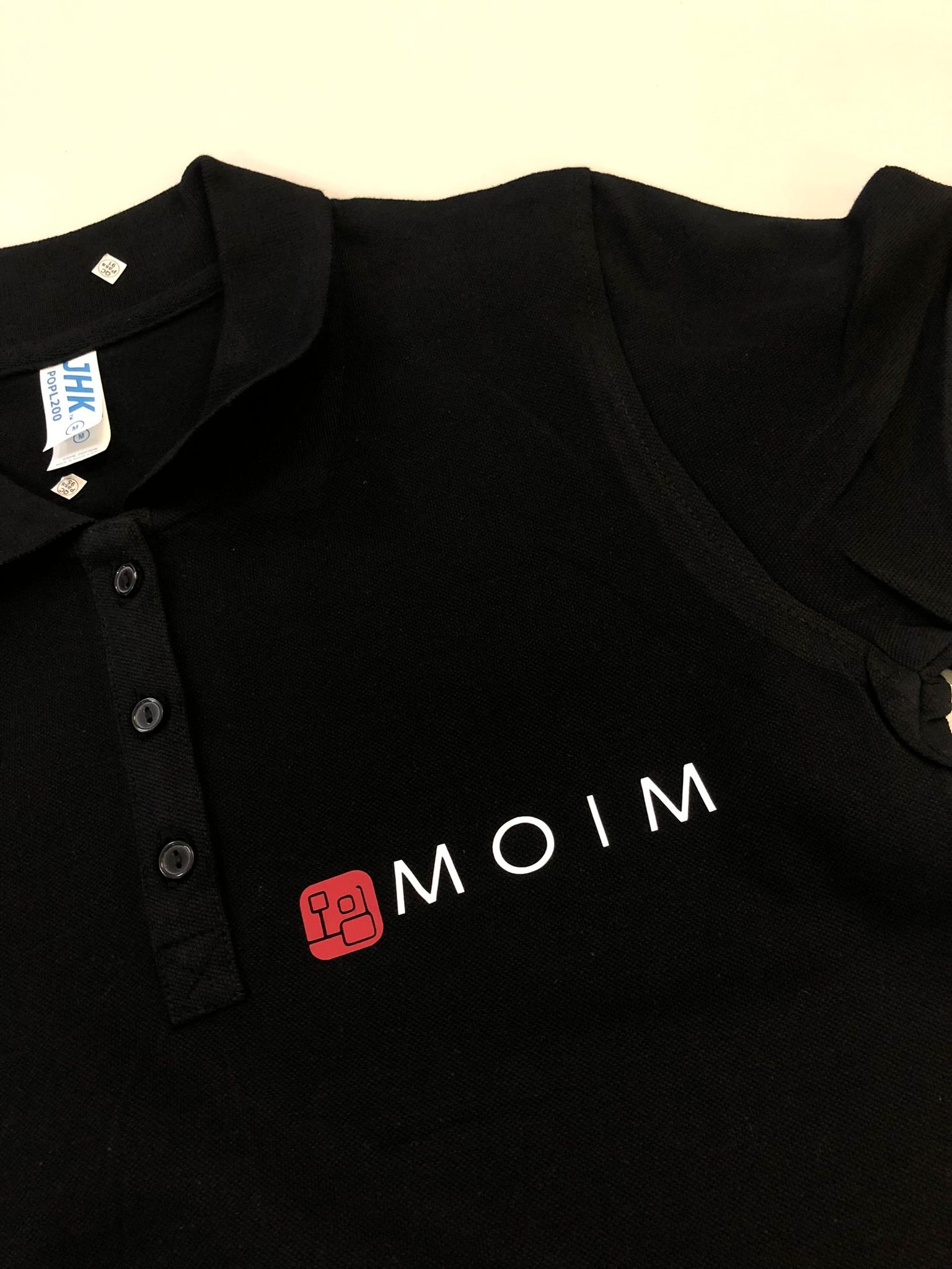 Czarna koszulka polo z białym napisem 'MOIM' i czerwonym logo. Widoczny dekolt, guziki i metka producenta. Materiał koszulki gładki, bez wyraźnej faktury.
