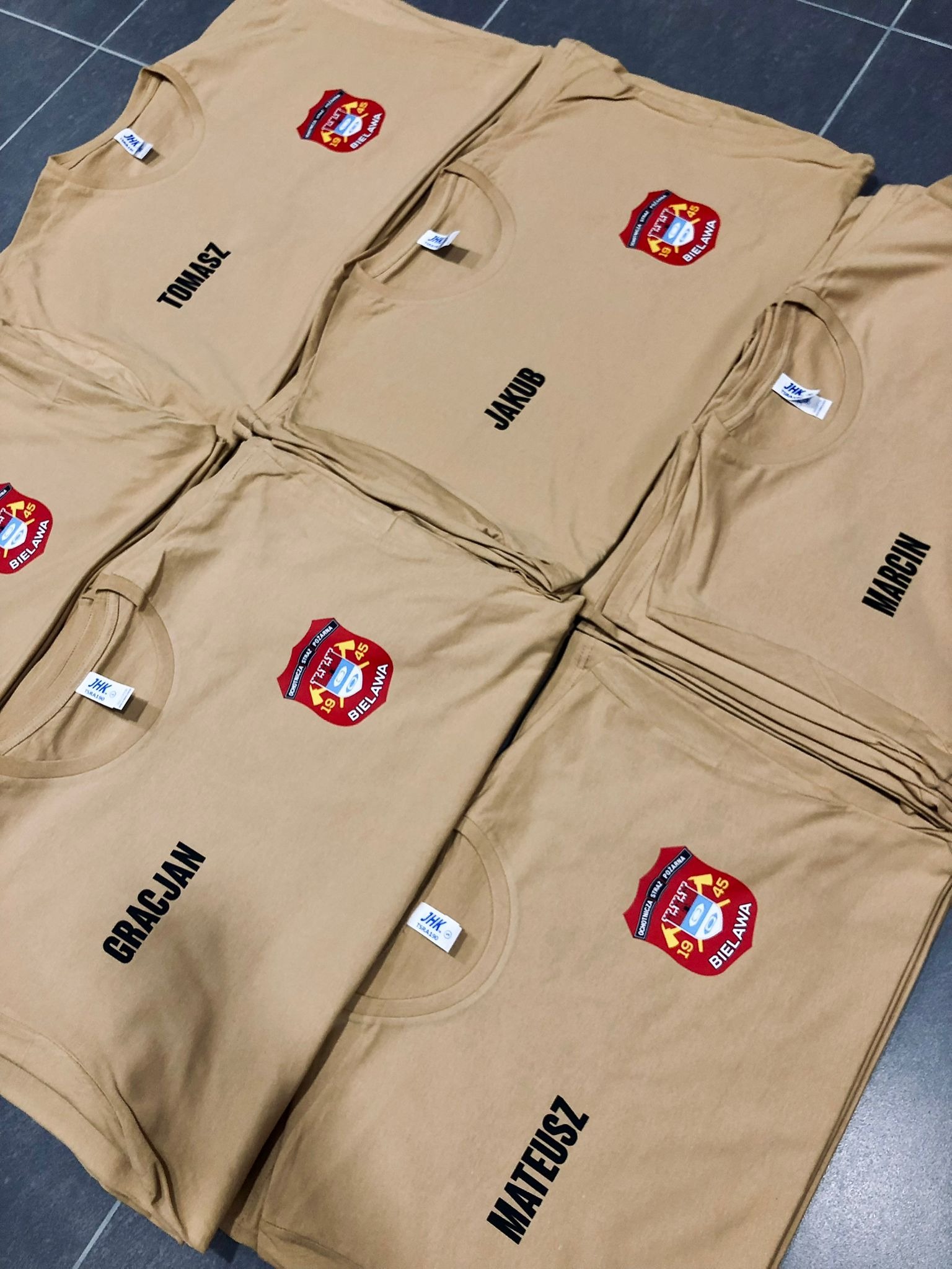 Beżowe koszulki z imionami i logiem OSP Bielawa, ułożone na podłodze. Nadruki wykonane czarną czcionką, logo w kolorze czerwonym i niebieskim. Widoczne metki JHK.
