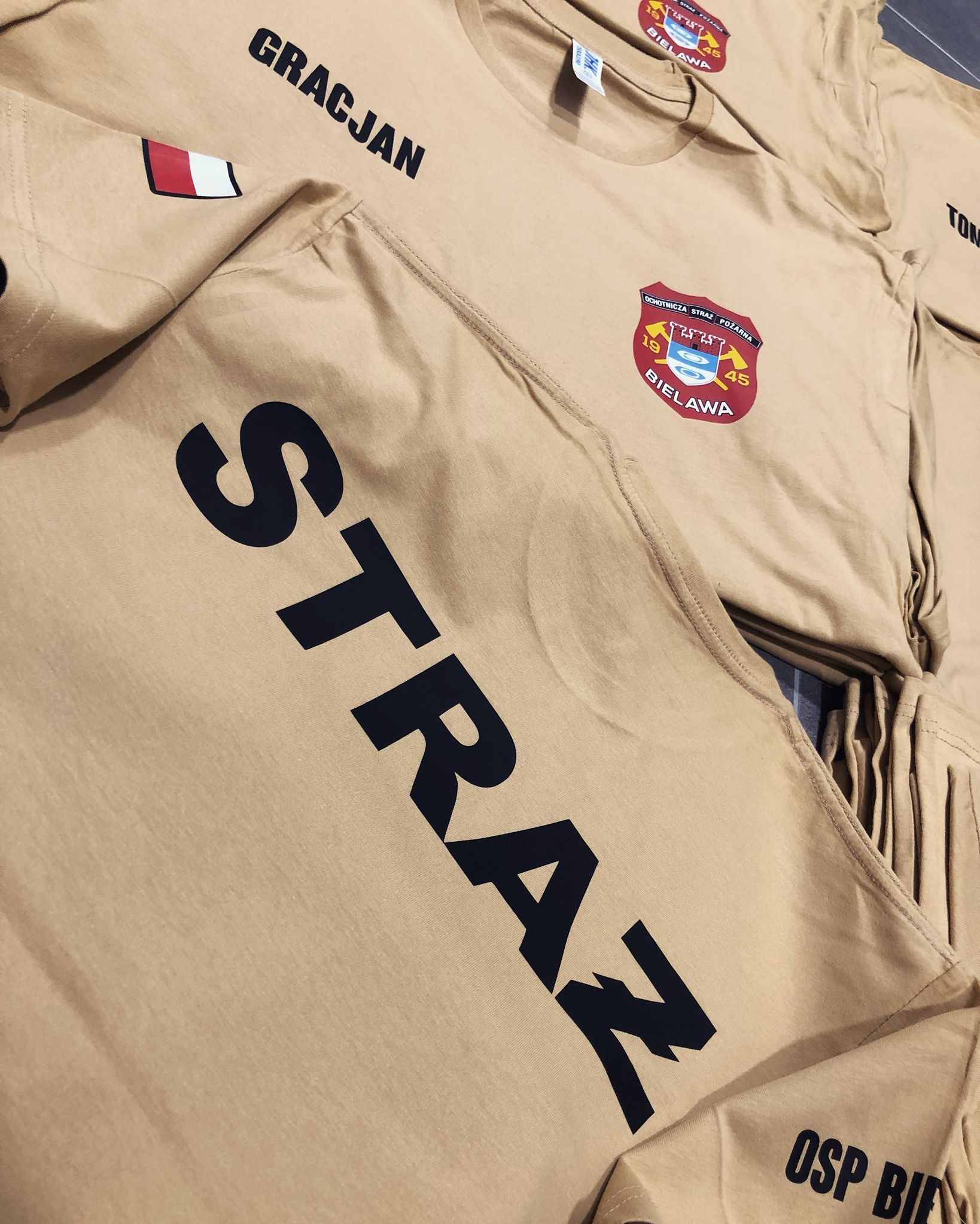 Beżowe koszulki z nadrukiem 'STRAŻ' i logiem OSP Bielawa, wykonane w Świebodzicach. Widoczne imiona i nazwy jednostek strażackich. Detal: naszywka z flagą.