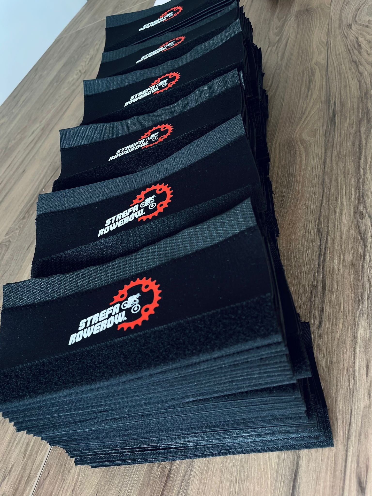 Stos czarnych, tekstylnych opasek z logo 'Strefa Rowerowa' w kolorze białym i czerwonym, leżących na drewnianej powierzchni. Widoczne detale materiału i nadruku.