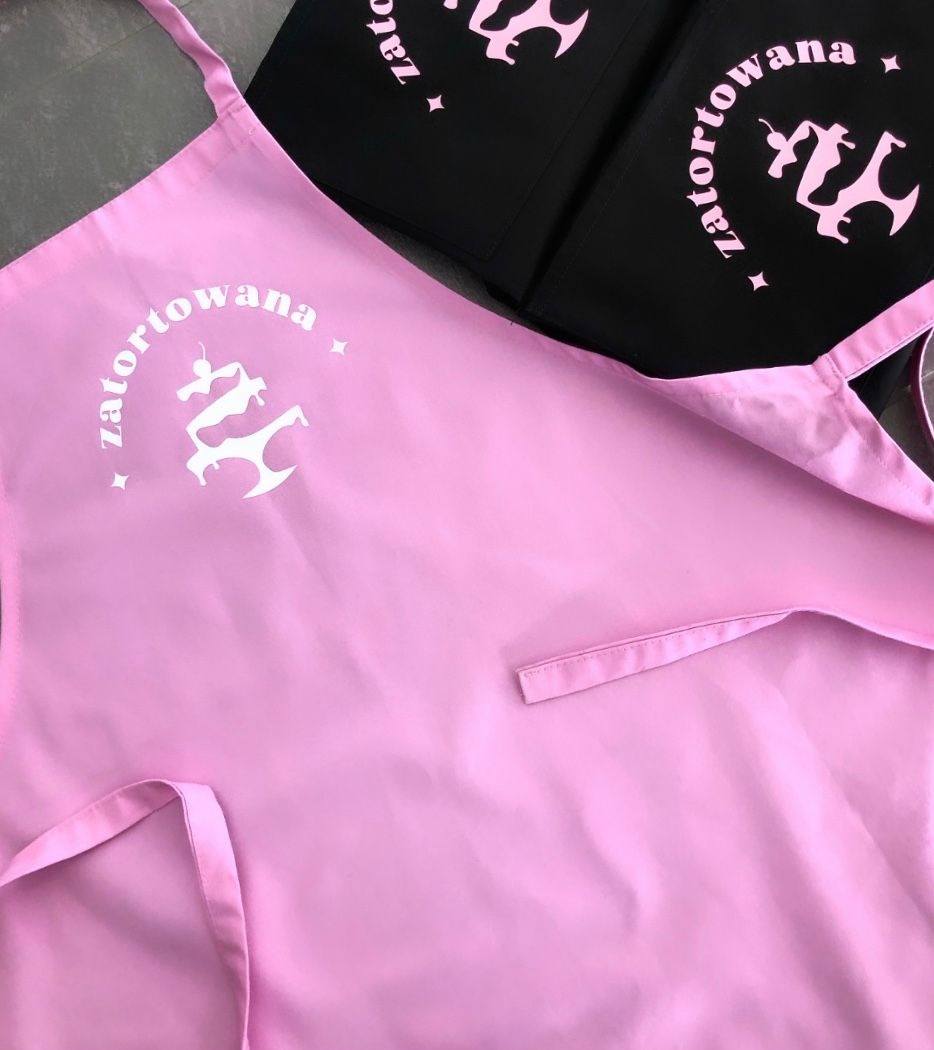Różowy fartuch z białym nadrukiem 'zatortowana' i logo, obok czarny fartuch z różowym nadrukiem, widoczne detale wiązań i kieszeni.