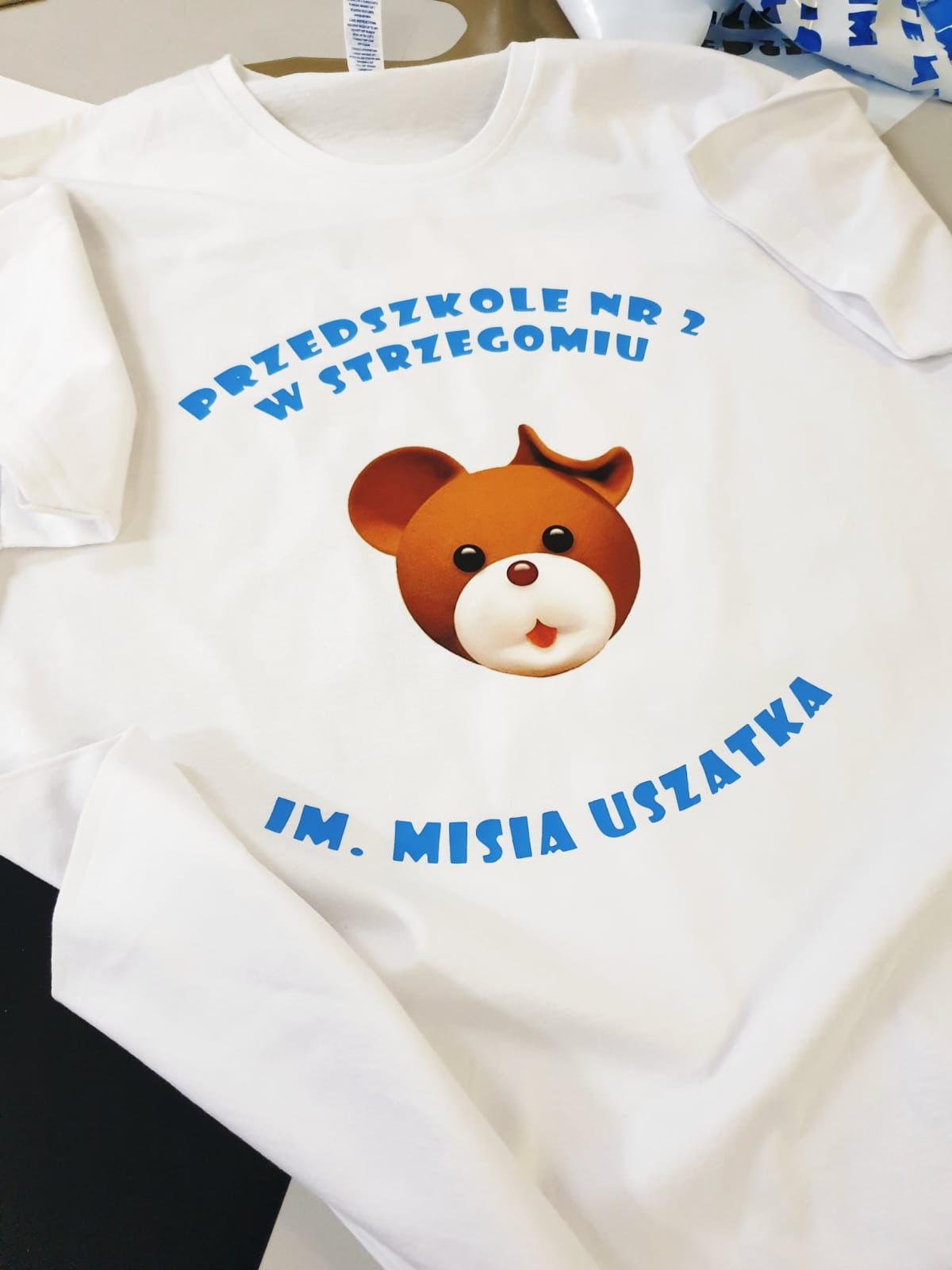 Biała koszulka z niebieskim nadrukiem: 'Przedszkole nr 2 w Strzegomiu Im. Misia Uszatka' i brązową grafiką misia, leżąca na stole.