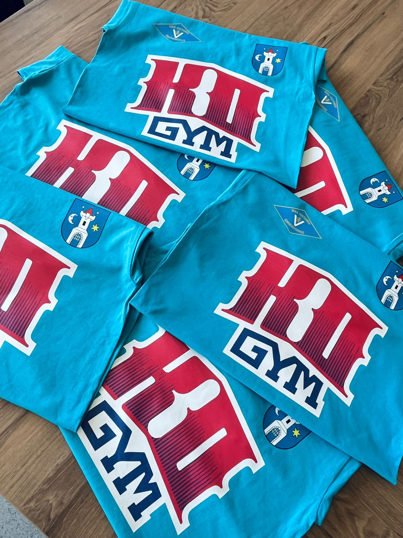 Stos turkusowych koszulek z czerwono-białym nadrukiem 'HIT GYM' i herbem Świebodzic, leżących na drewnianym podłożu. Detaliczny widok na odzież sportową.