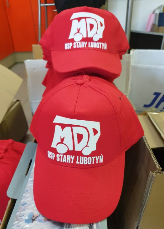 Czerwone czapki z białym nadrukiem 'MDP OSP Stary Lubotyń' ułożone jedna na drugiej. Widoczne detale nadruku i faktura materiału czapek.