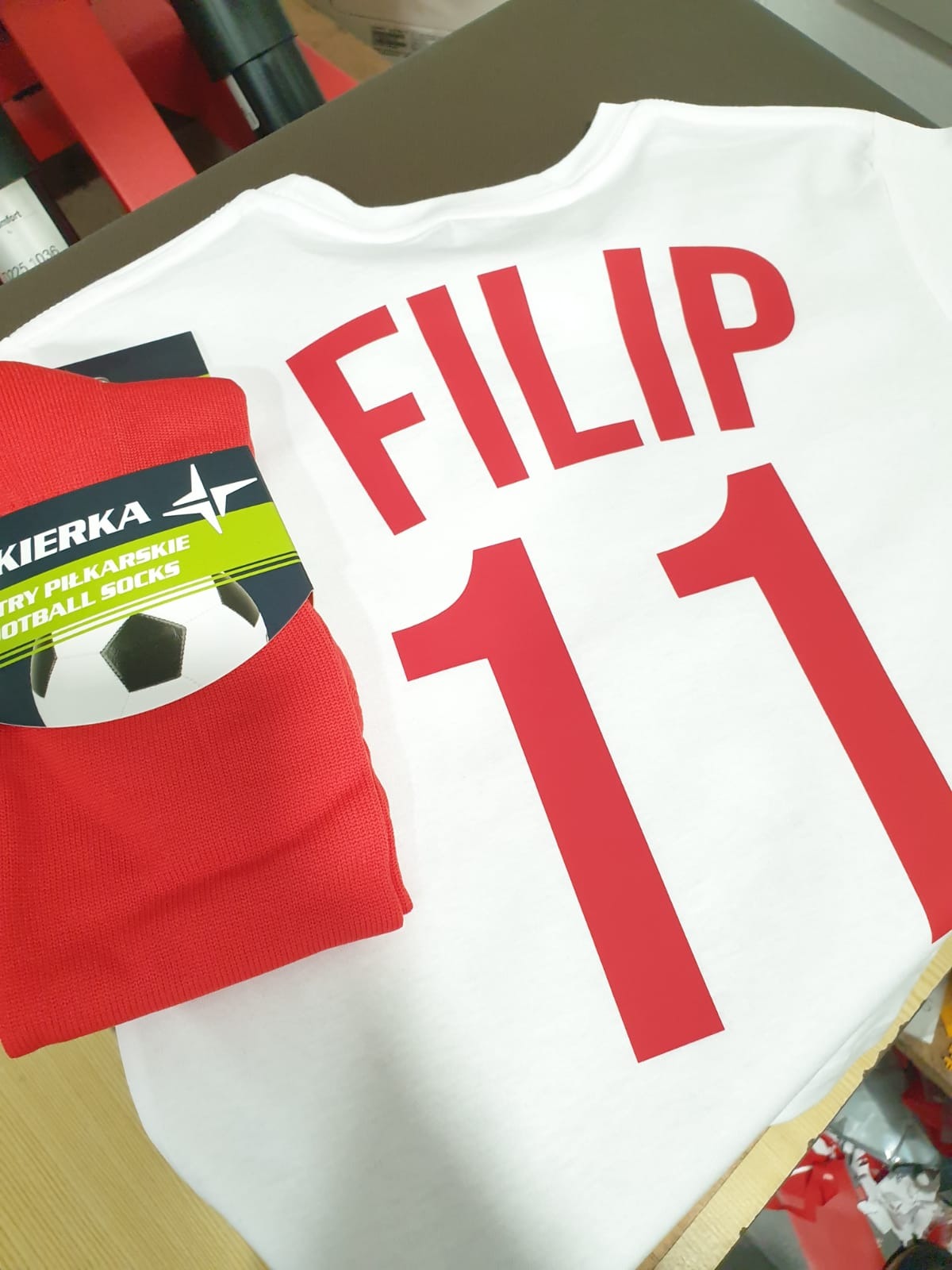 Biała koszulka z czerwonym nadrukiem 'FILIP 11' leży obok czerwonych, zapakowanych skarpetek piłkarskich, tworząc komplet kibica.