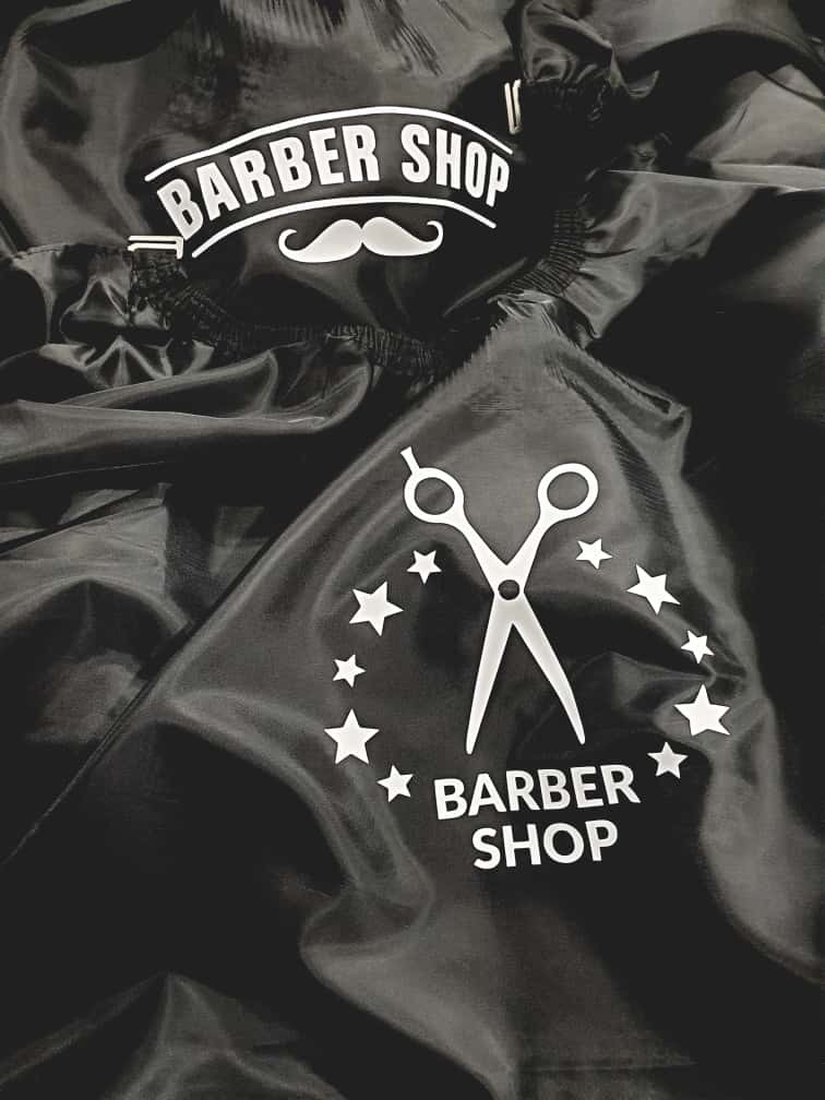 Czarna peleryna fryzjerska z białym nadrukiem: nożyczki, gwiazdy i napis 'Barber Shop' z wąsami. Materiał lekko pognieciony, widoczne detale wykończenia.