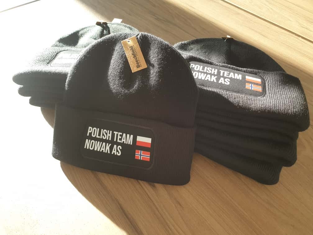 Czarne, dziergane czapki z napisem 'POLISH TEAM NOWAK AS' i flagami Polski i Norwegii, ułożone na drewnianej powierzchni w słonecznym świetle.