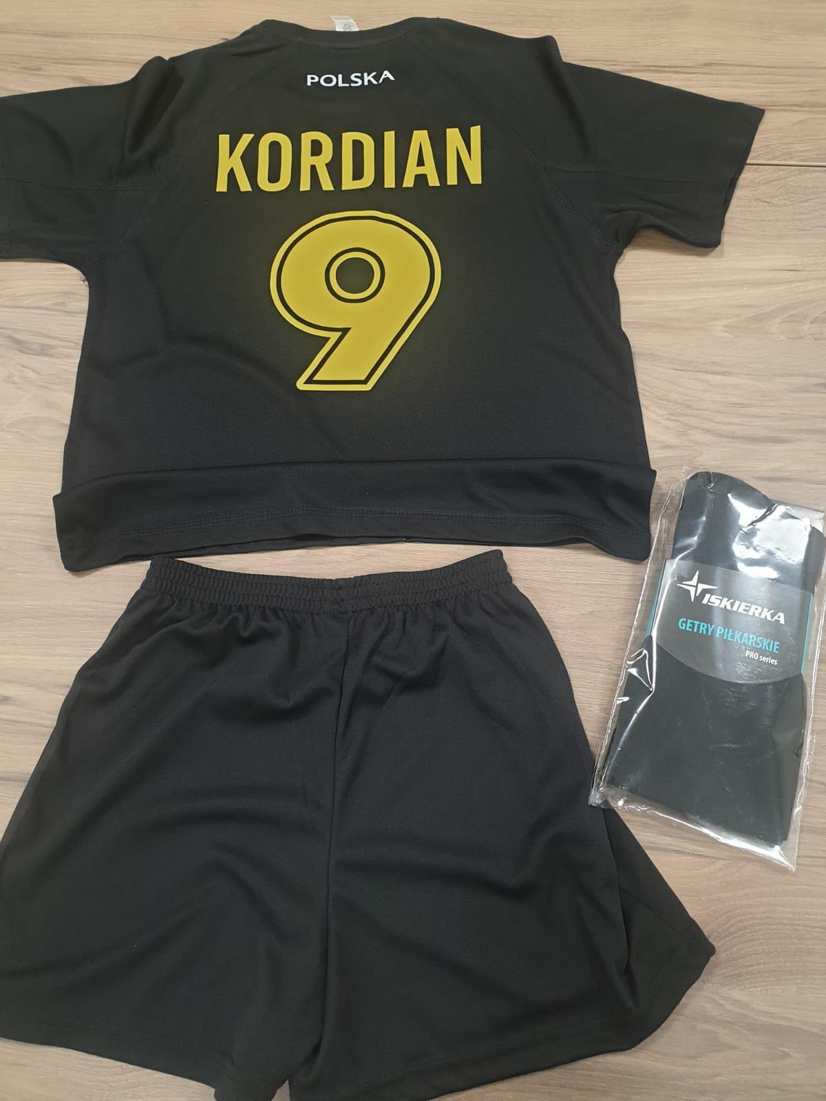 Czarny komplet sportowy: koszulka z napisem 'POLSKA', imieniem 'KORDIAN' i numerem 9, spodenki oraz zapakowane getry piłkarskie 'ISKIERKA'.