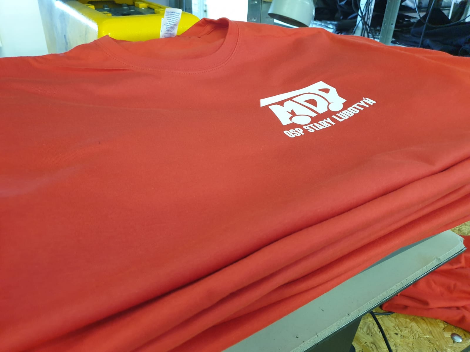 Czerwona koszulka z białym nadrukiem logo MDP OSP Stary Lubotyń, leżąca na stole. Widoczne fałdy materiału i fragment maszyny do nadruków.