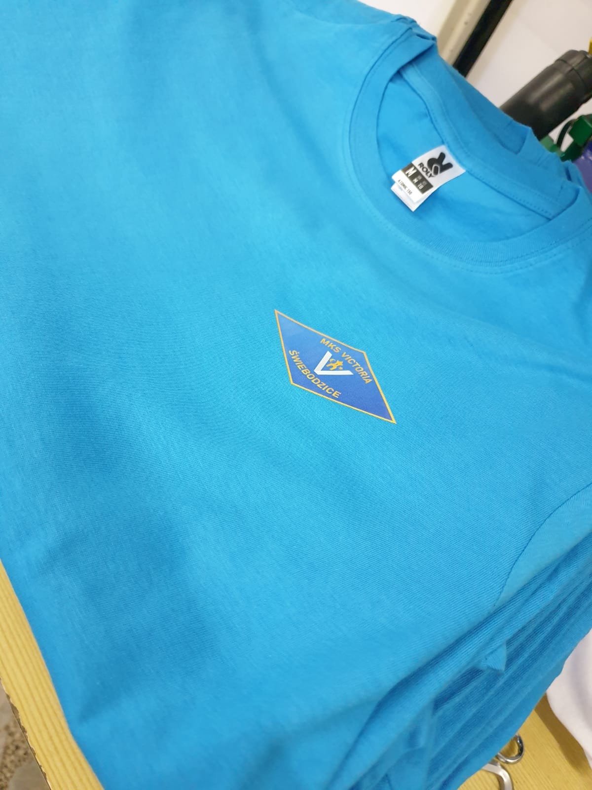 Jasnoniebieska koszulka z nadrukiem logo MKS Victoria Świebodzice. Widoczna metka producenta Roly. Ułożona w stosie z innymi koszulkami.
