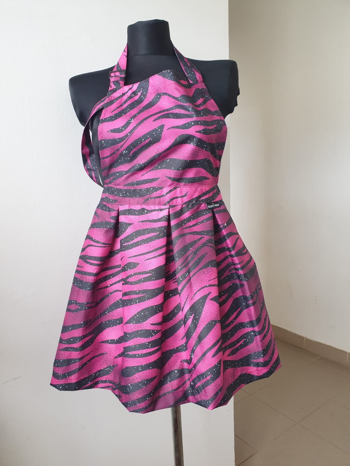 Różowo-czarny fartuch damski w zebrę na manekinie. Fartuch z plisowaną spódnicą i regulowanym paskiem na szyję. Widoczna metka 'BEAUTY-WEAR'.