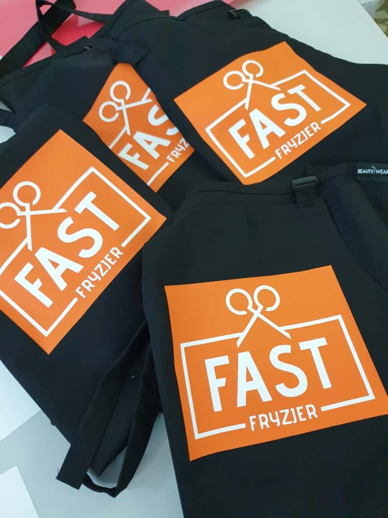 Czarne fartuchy fryzjerskie z pomarańczowym logo 'FAST Fryzjer' i ikoną nożyczek. Fartuchy ułożone warstwowo, widoczne paski do zawiązywania.