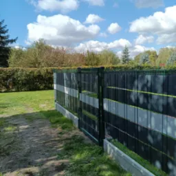 Zielone ogrodzenie panelowe z szarymi przesłonami na betonowej podmurówce, widok z boku na tle zieleni i błękitnego nieba z chmurami. Ukazany fragment ścieżki.