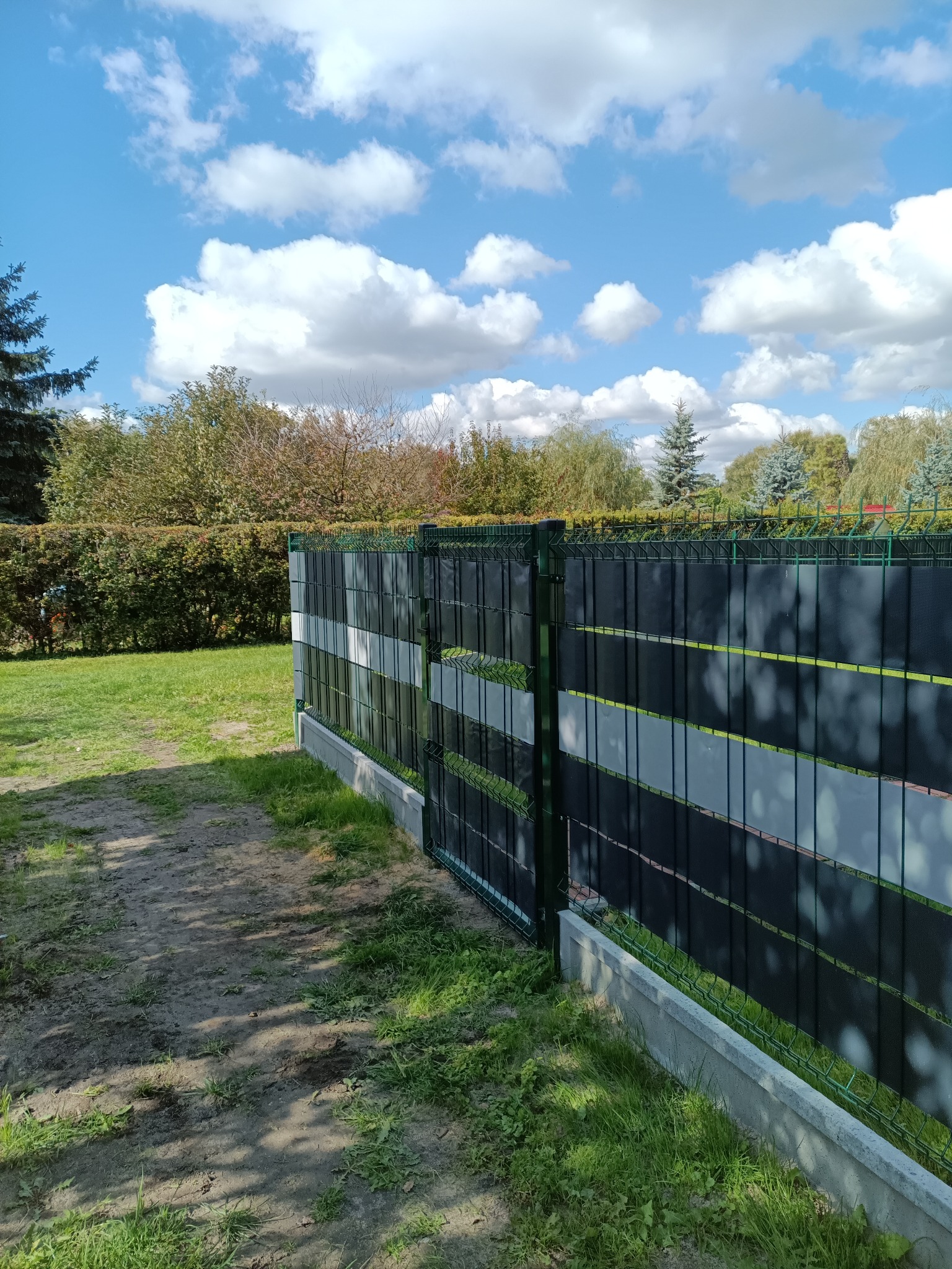 Zielone ogrodzenie panelowe z szarymi przesłonami na betonowej podmurówce, widok z boku na tle zieleni i błękitnego nieba z chmurami. Ukazany fragment ścieżki.