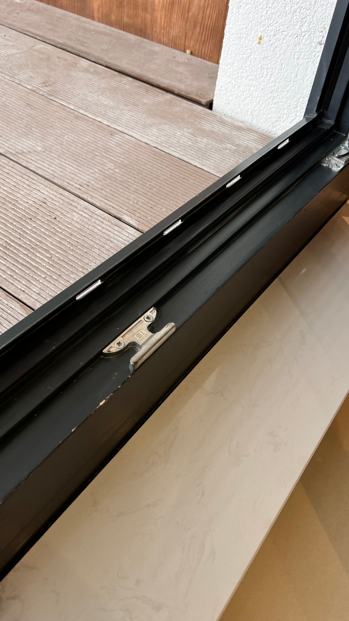 Szczegółowe ujęcie czarnej ramy okiennej z widocznym mechanizmem zamknięcia, fragmentem drewnianego tarasu i jasną, marmurkową podłogą. Widoczne zabrudzenia.