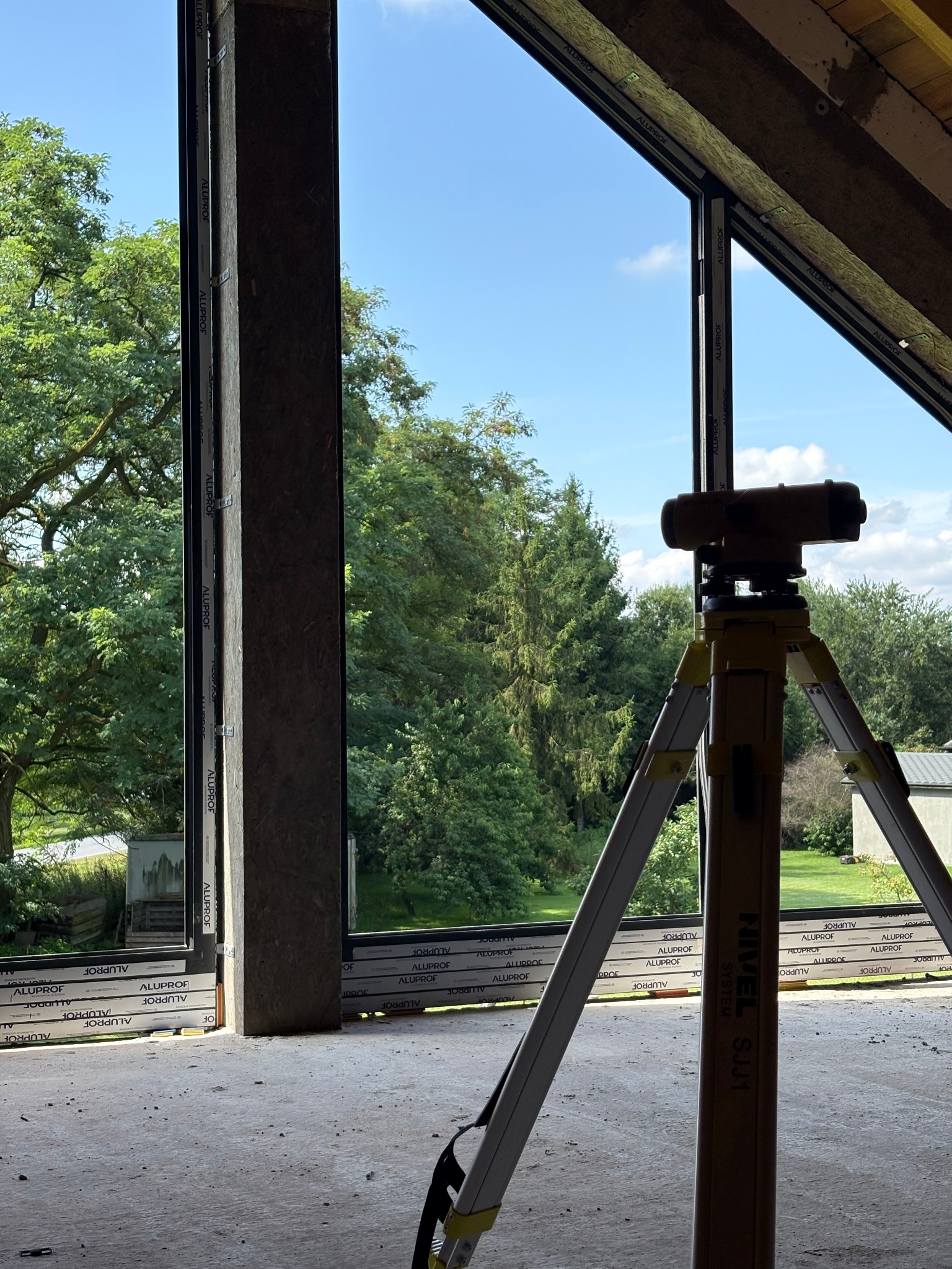 Montaż okna marki Aluprof w surowym wnętrzu poddasza. Na pierwszym planie niwelator na statywie. Widok na zewnątrz przez okno z zielenią drzew i błękitnym niebem.