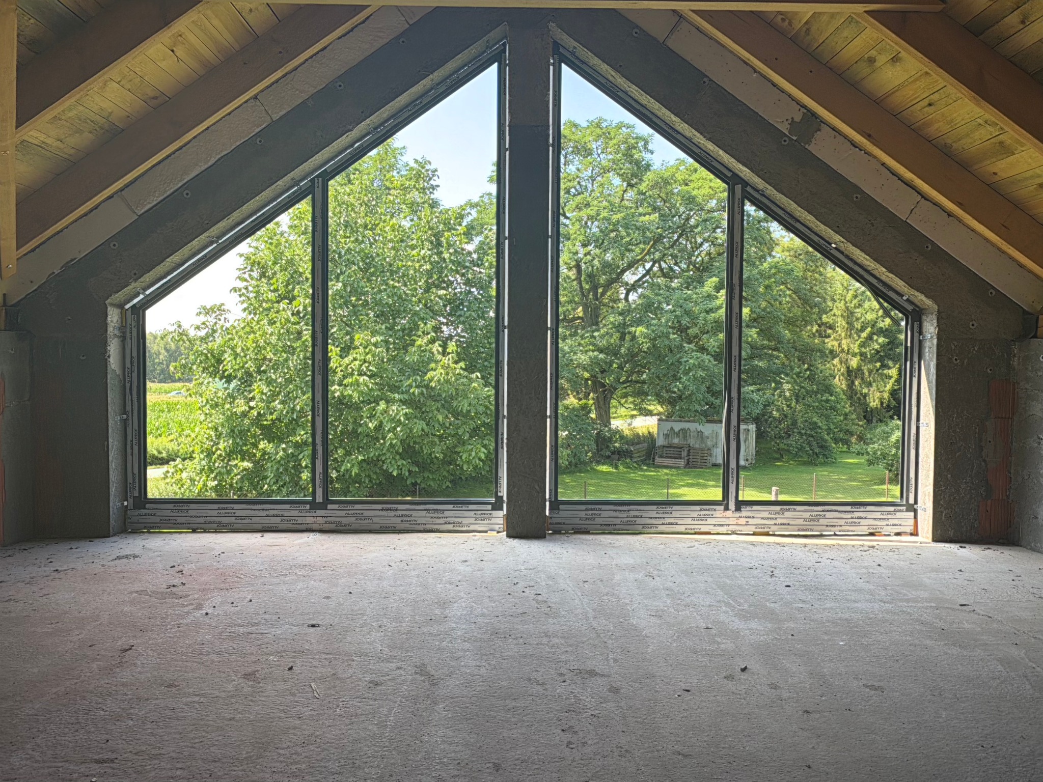 Trzy okna aluminiowe zamontowane w surowym stanie poddasza z drewnianą więźbą dachową. Widok na zieleń za oknem. Szare ramy okienne.