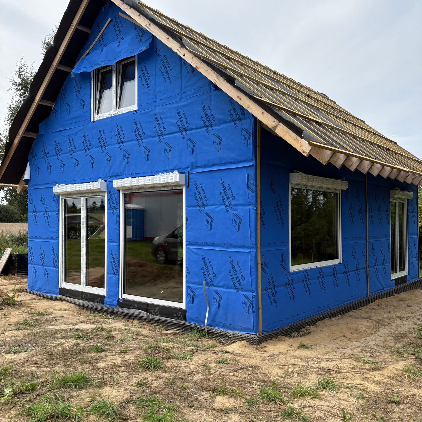 Dom w trakcie budowy, z zamontowanymi oknami PCV i niebieską membraną paroizolacyjną na ścianach. Widoczne drewniane elementy konstrukcji dachu.