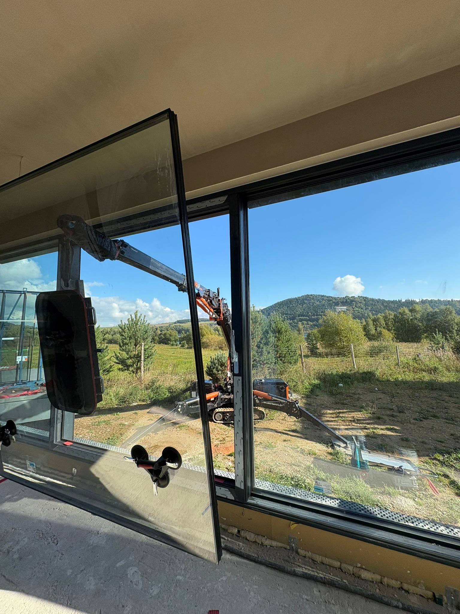 Montaż okna panoramicznego z użyciem dźwigu na tle malowniczego krajobrazu. Widoczne przyssawki do szkła i elementy konstrukcyjne.