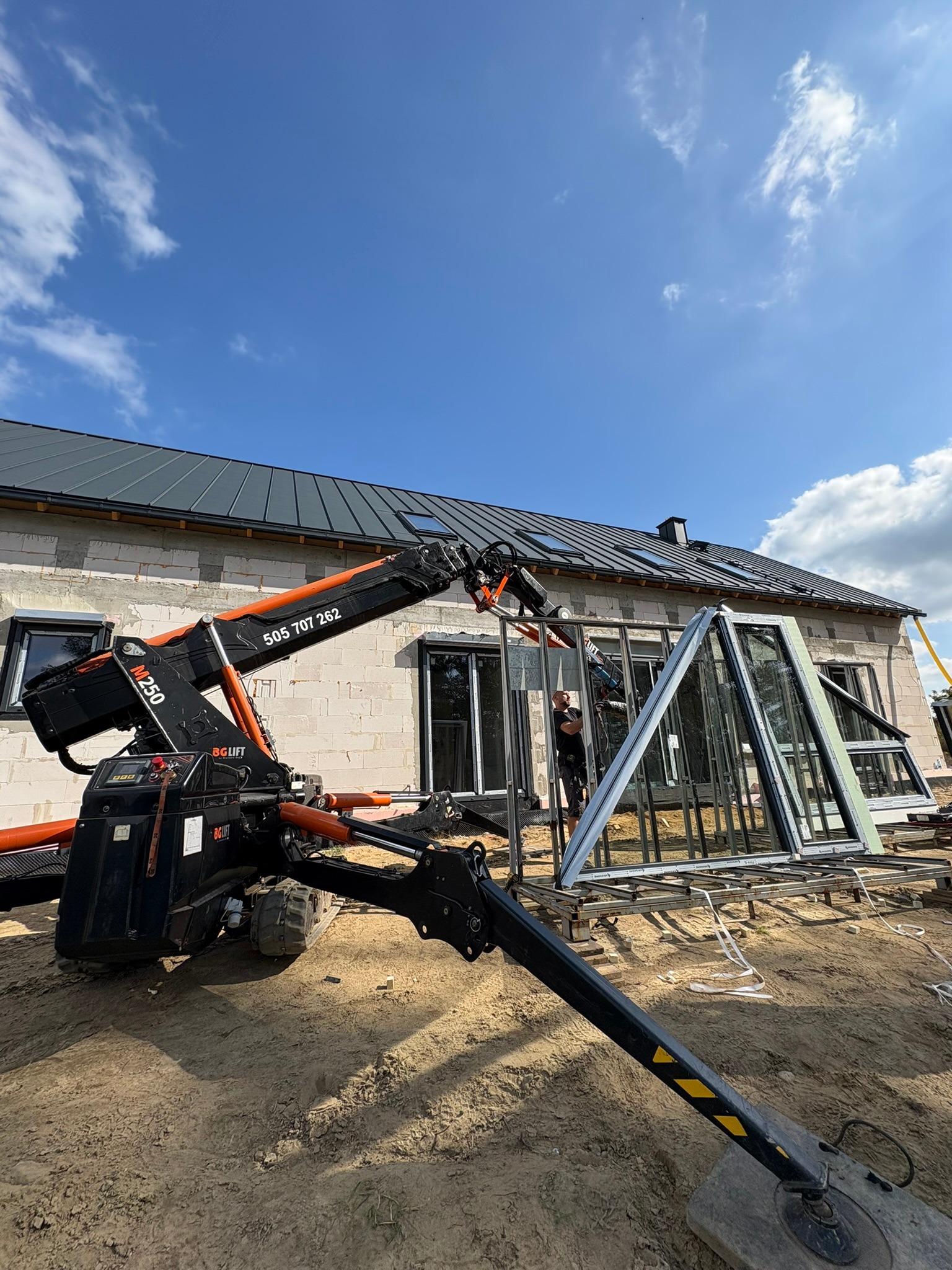 Montaż okien na nowym budynku za pomocą podnośnika. Widoczne okna aluminiowe, pracownik i fragment ściany z oknami. Błękitne niebo w tle.