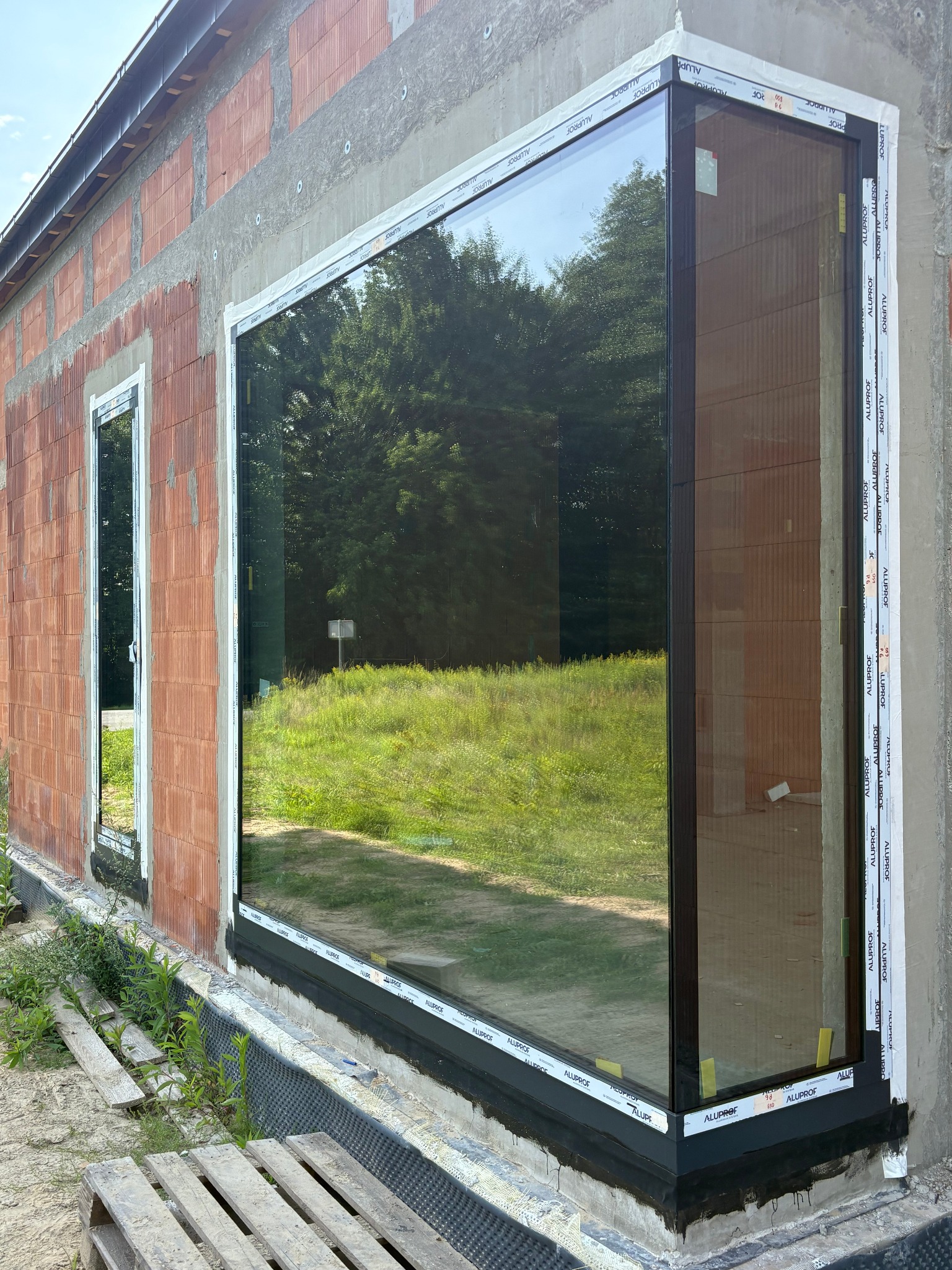 Montaż dużego okna z brązową ramą w surowym murze. Widoczne detale montażowe i folia ochronna. Okno odbija krajobraz z zielenią.