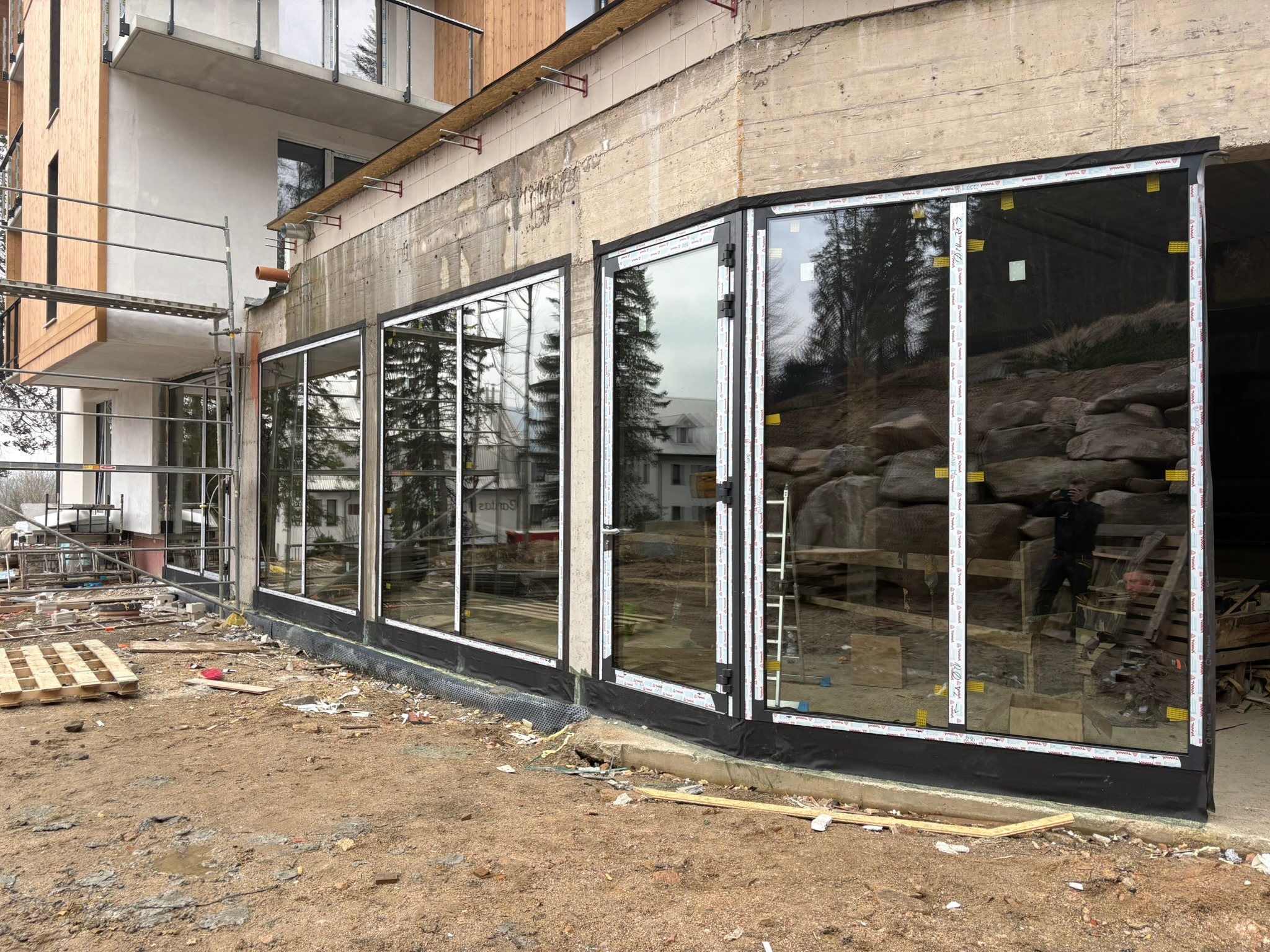 Montaż okien z czarną ramą w Kobierzycach. Widoczne zabezpieczenia na szybach, surowy stan budynku, ziemia. W tle odbicie budynku i zieleni w oknach.