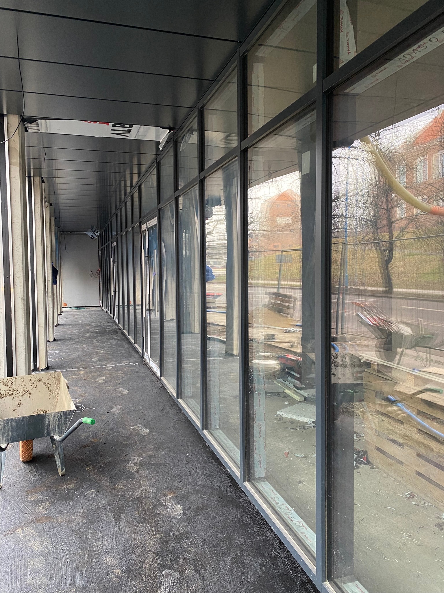 Długi korytarz z zamontowanymi oknami aluminiowymi, widoczny wózek budowlany i ślady na podłodze, sugerujące trwające prace wykończeniowe.