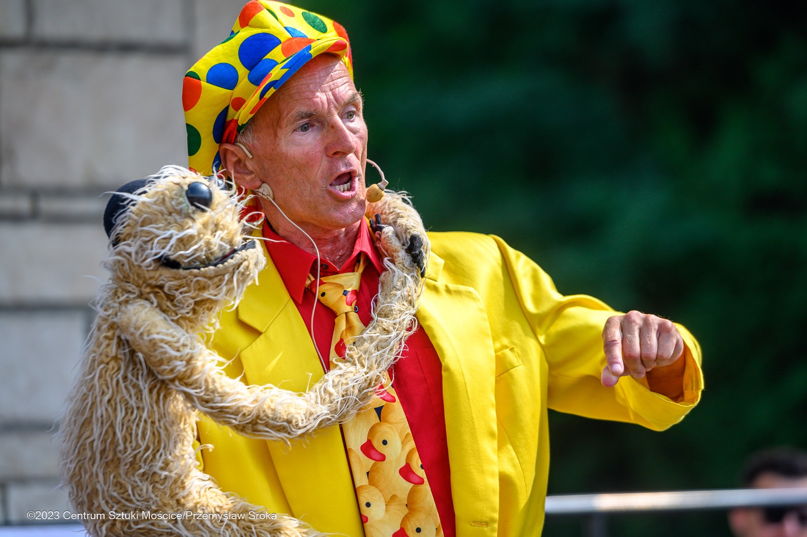 Animator w żółtym kostiumie i czapce w kropki, z lalką szmacianą, w trakcie występu. Wyrazista mimika, dynamiczna poza. Tło zielone, rozmyte.
