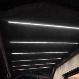 ElektroMax Piotr Jakubek - Drewniany sufit z równomiernie rozmieszczonymi, podłużnymi lampami LED, oświetlającymi przestrzeń w ciemnym otoczeniu. Widoczne elementy konstrukcyjne dachu.