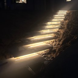 ElektroMax Piotr Jakubek - Oświetlone schody zewnętrzne prowadzące w górę, z ciepłym światłem podkreślającym fakturę kamienia i ziemi. Nocne ujęcie z niskim kątem, akcentuje nowoczesny design oświetlenia.